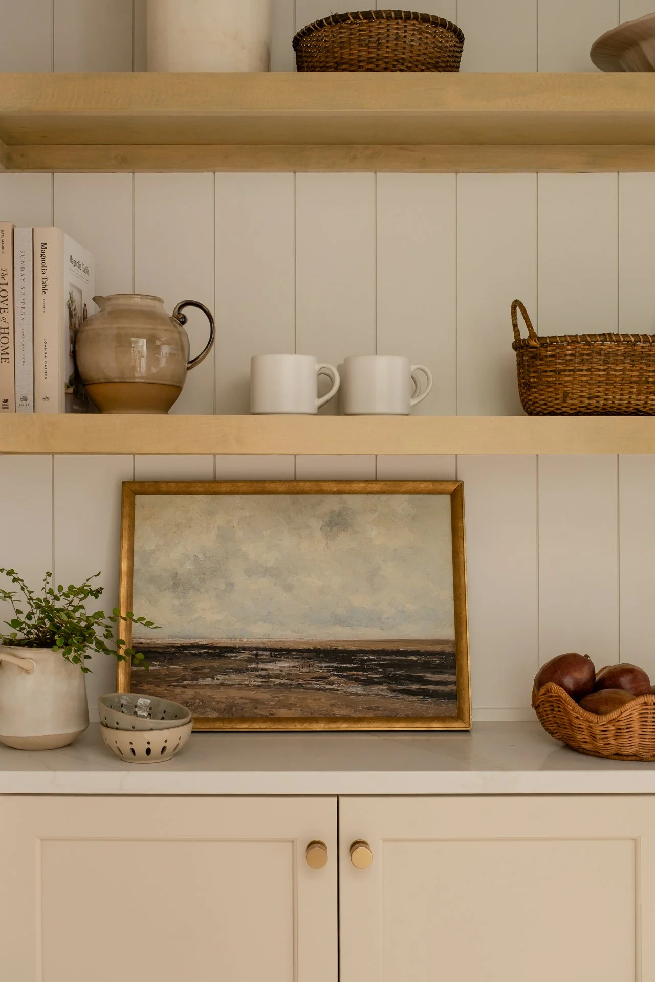 Open shelving with books, a ceramic pitcher, white mugs, a wicker basket, a framed landscape painting, a potted plant, and a bowl of onions on a white cabinet.