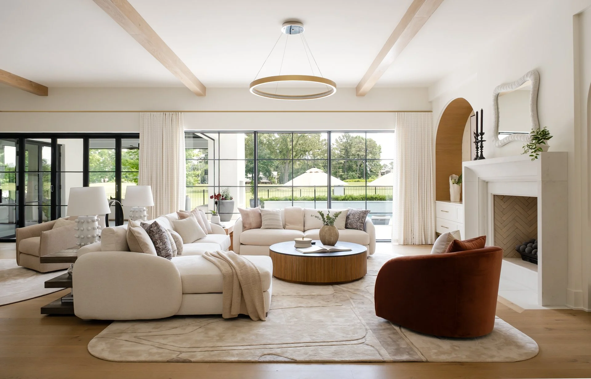 Bright living room with beige sofas, a round wooden coffee table, armchair, large windows with white curtains, fireplace, and minimalist decor.