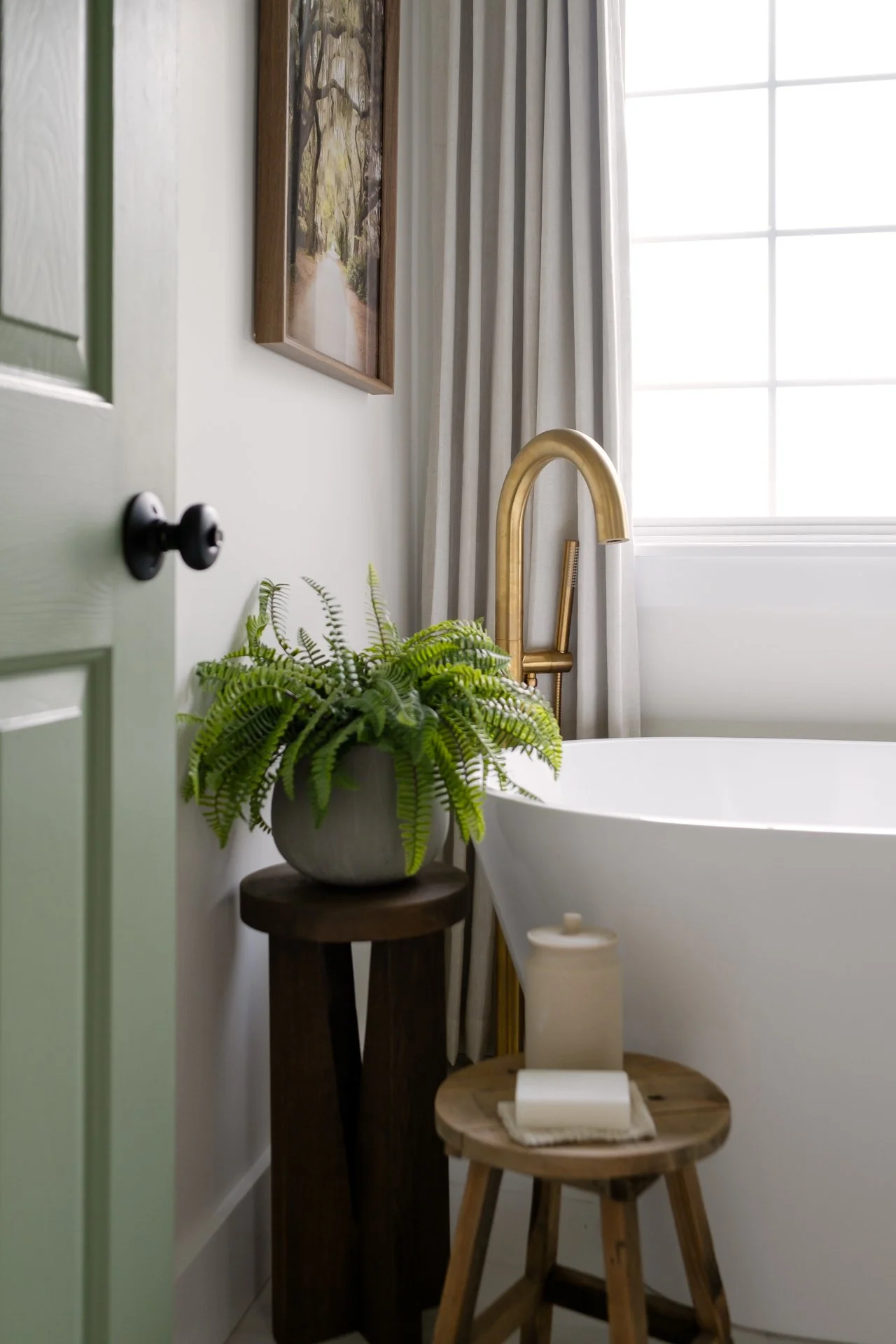 A bathroom with a white bathtub, gold faucet, green door, potted fern plant on a wooden stand, window with light curtains, and wall decor.
