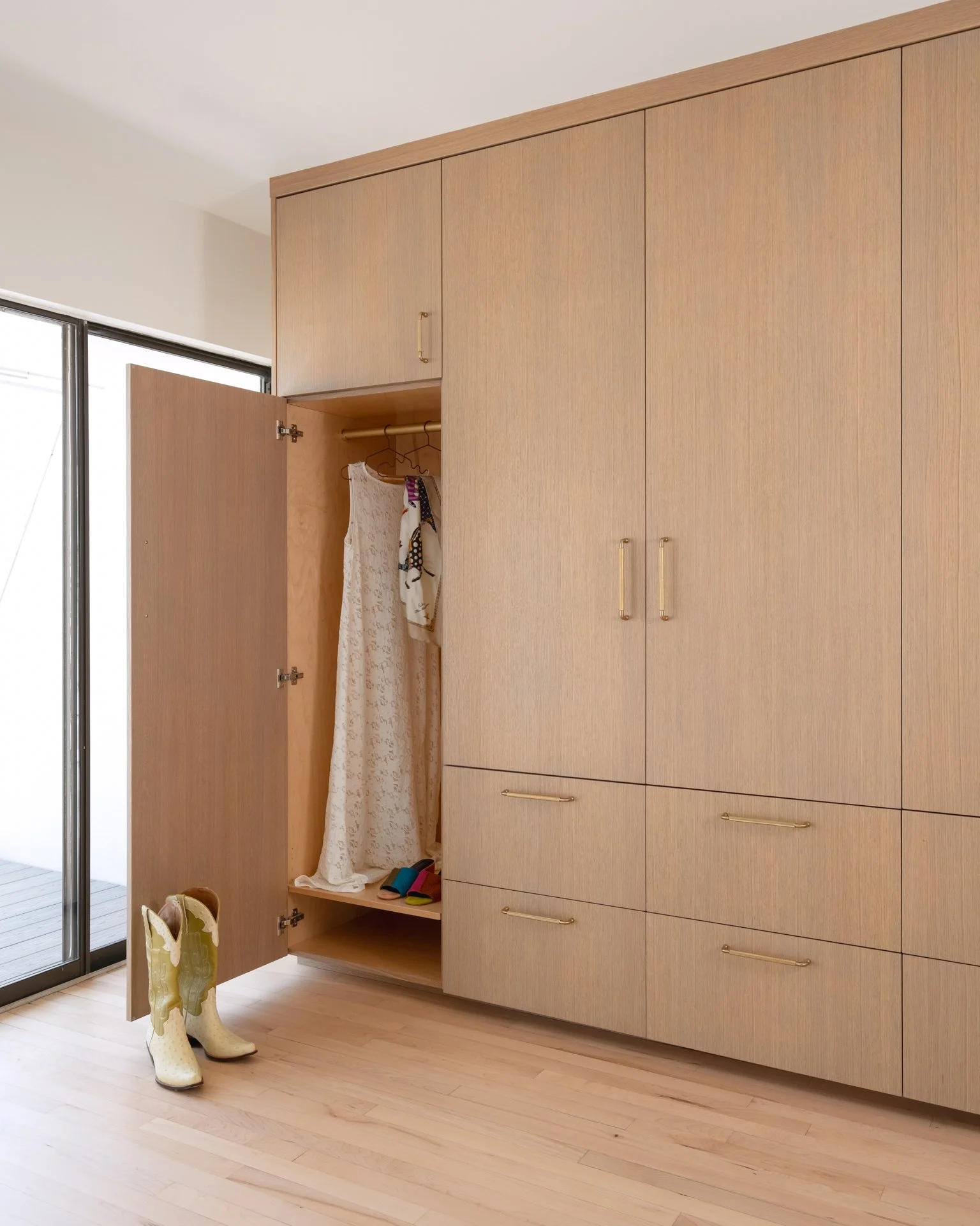 Open wooden wardrobe with a dress hanging inside, a pair of cowboy boots on the floor next to it, and a sliding glass door in the background.