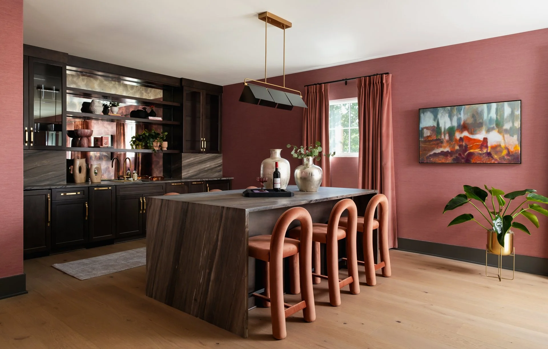 Modern kitchen with pink walls, dark wood cabinets, a marble backsplash, a wooden island with three pink chairs, a window with pink curtains, a hanging black and gold light fixture, decorative vases on the island, a potted plant, and a landscape pain
