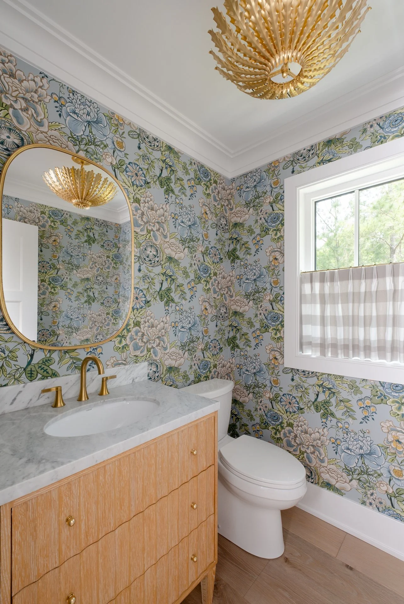 A bathroom with floral wallpaper, a gold-framed mirror, a window with a white and beige striped blind, a light wood vanity with a marble countertop, a white toilet, and a gold decorative ceiling light fixture.