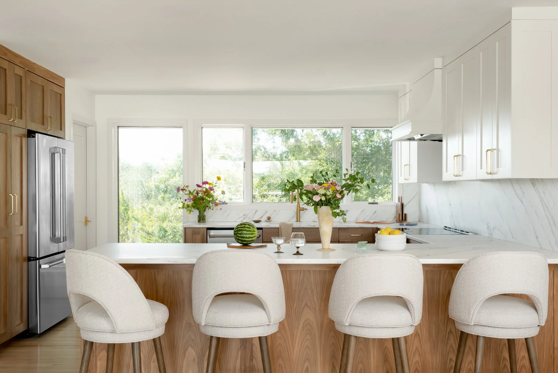 Bright kitchen with white and wood cabinets, marble backsplash and island, large windows with greenery outside, decorative vases with flowers, watermelon on the island, and beige barstools.