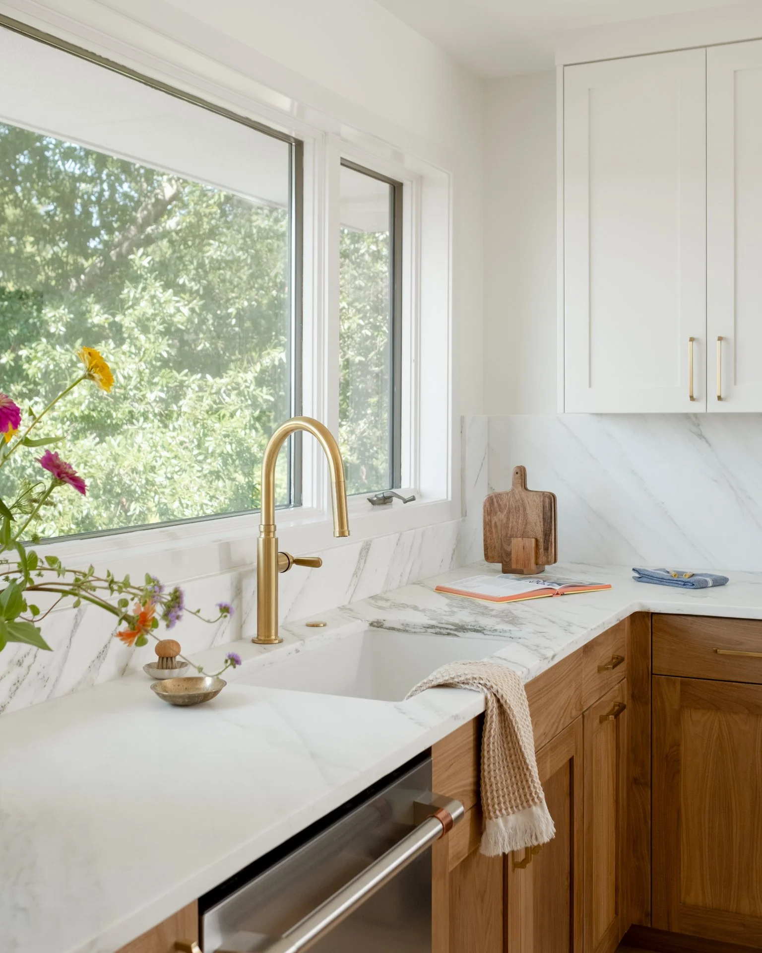 Modern kitchen with white marble countertops, wooden cabinets, and a large window showing green trees outside.