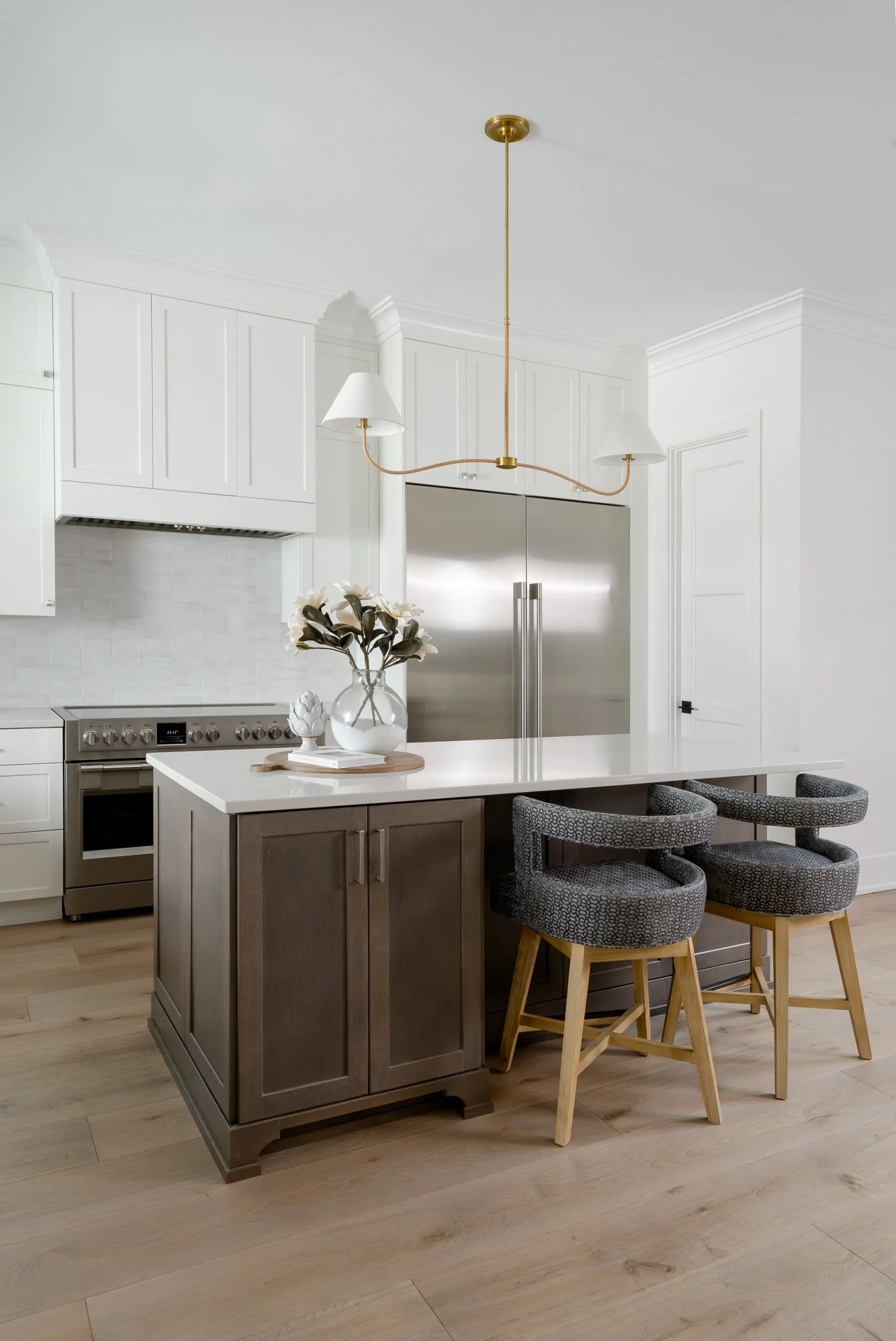 A modern kitchen with white cabinets, stainless steel refrigerator, and a kitchen island with a dark wood base and white countertop. Two patterned bar stools with wooden legs are positioned at the island. A gold chandelier with two white lampshades h