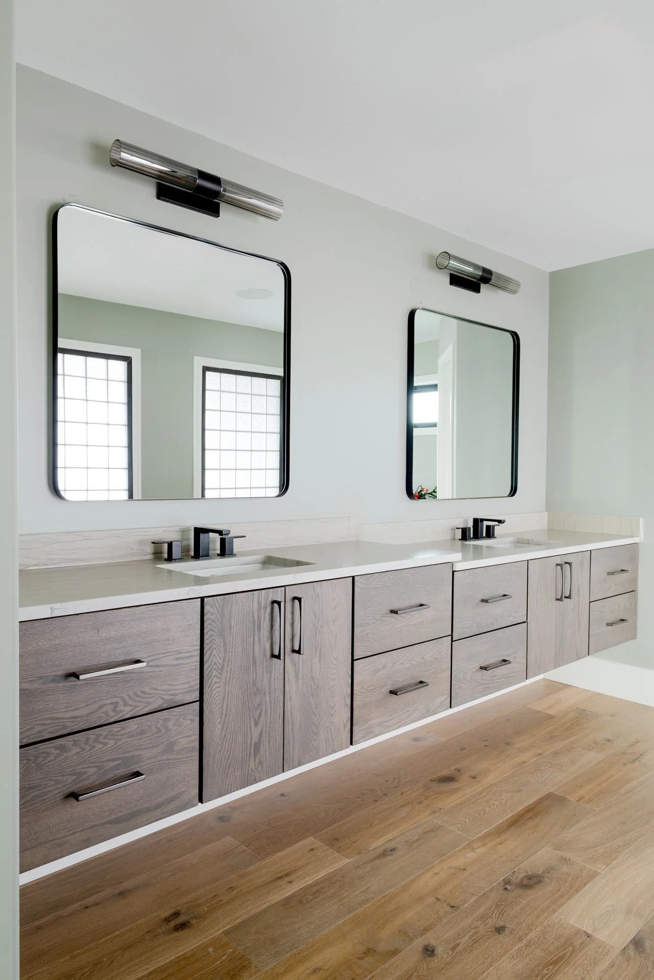 Double bathroom vanity with two mirrors and two black faucets, set against a white wall with wooden flooring.