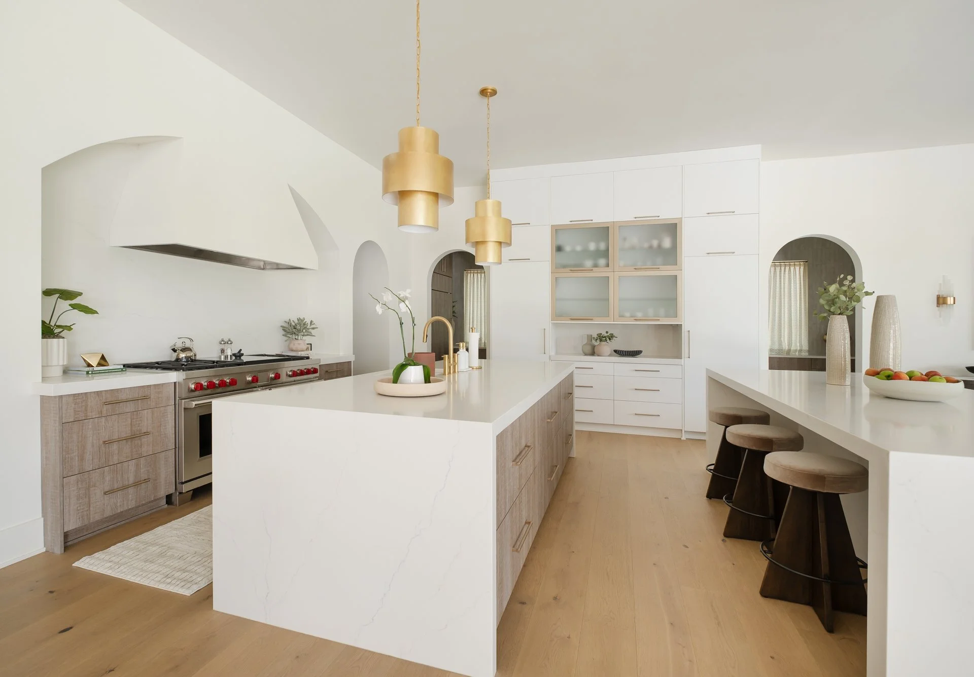 Modern white and wood kitchen with island, bar stools, gold pendant lights, plants, and stainless steel appliances
