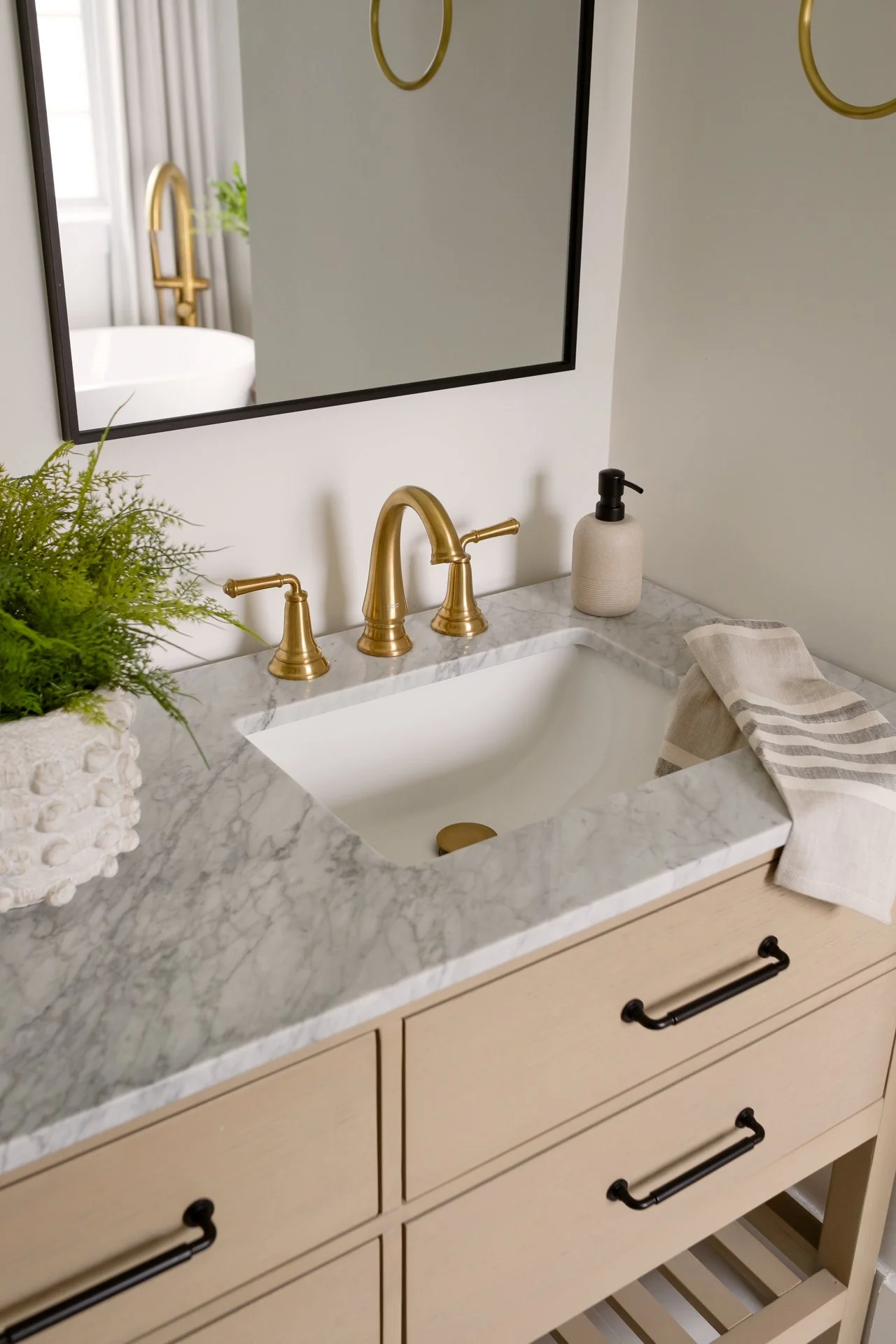 Bathroom sink with a marble countertop, gold faucet, a plant in a textured pot, black pump soap dispenser, striped towel, and a mirror reflecting a bathtub with a gold faucet and a window.