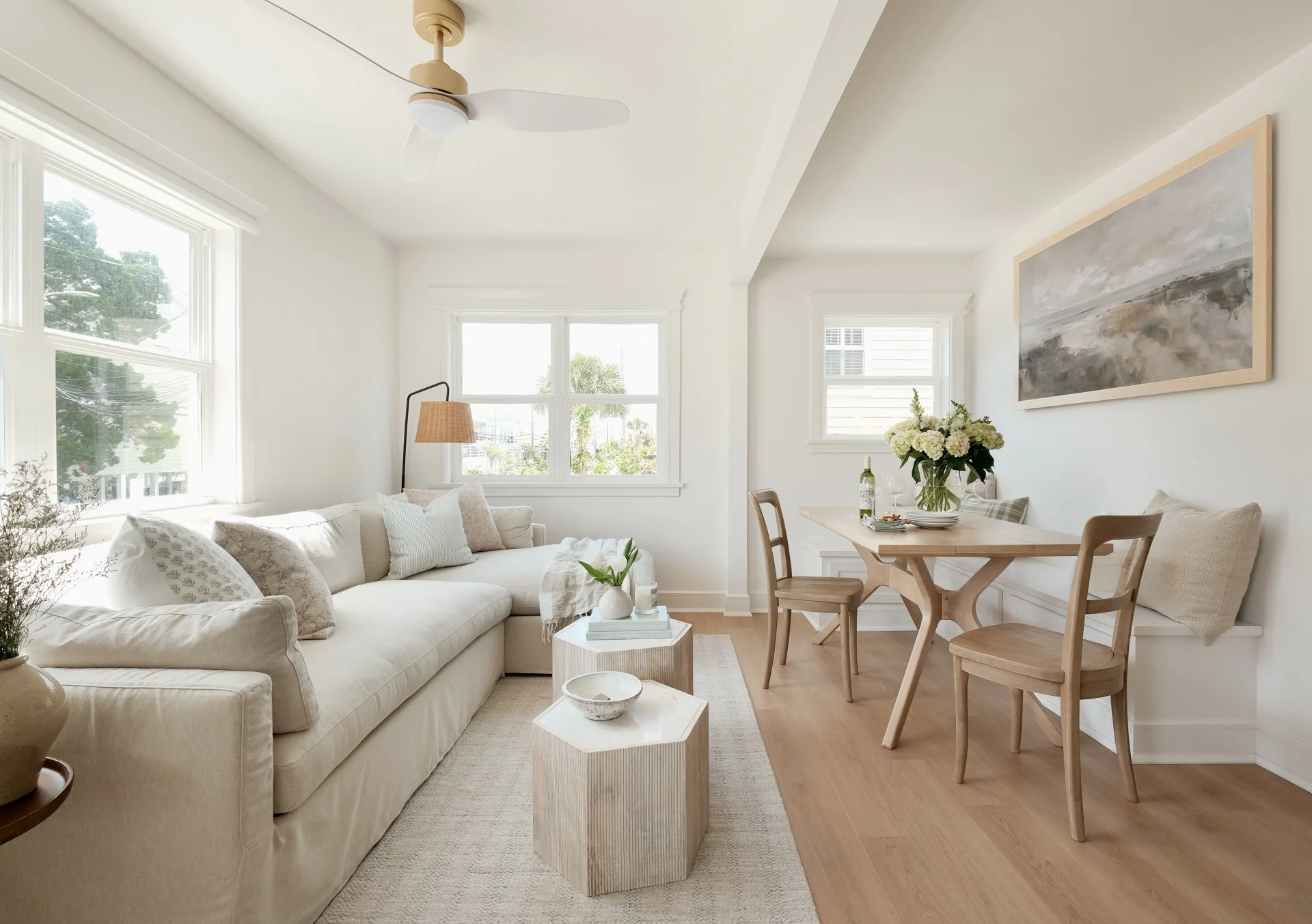 Bright living room with white walls and furniture, wooden dining table with chairs, large windows, and minimal decor.