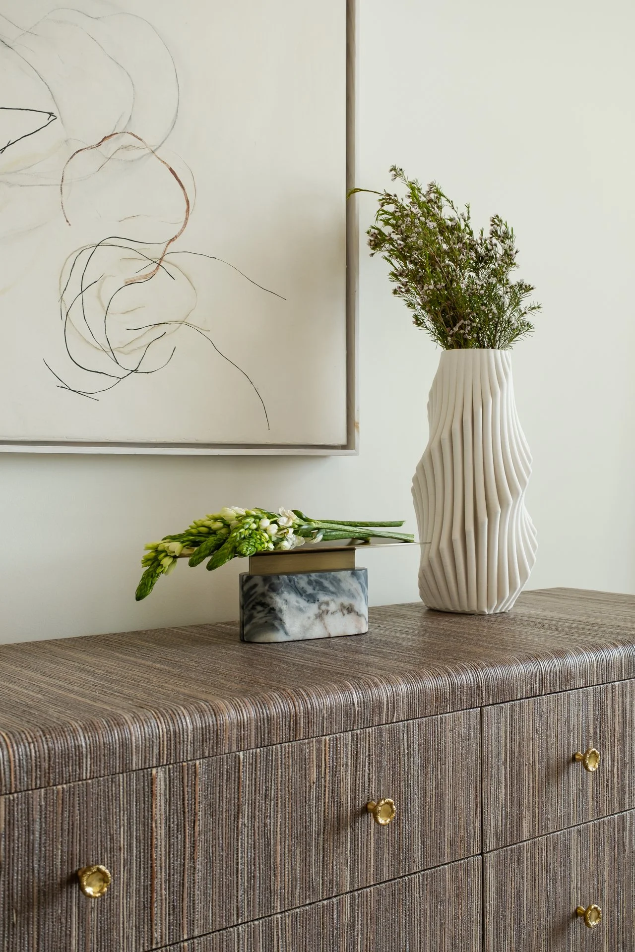 A wooden sideboard with a marble box, a flower arrangement, and a tall textured white vase with greenery, against a white wall with an abstract line art painting.