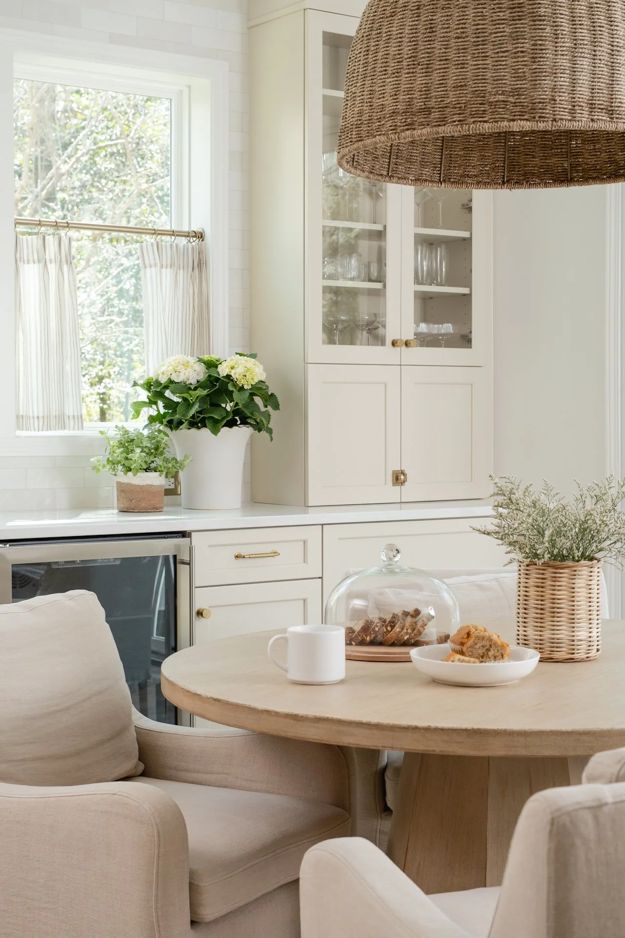A cozy kitchen corner with a round wooden table, beige armchairs, a white cabinet with glass doors, and a window with striped curtains. Decor includes potted plants, a cake stand with cookies, and a basket with greenery.