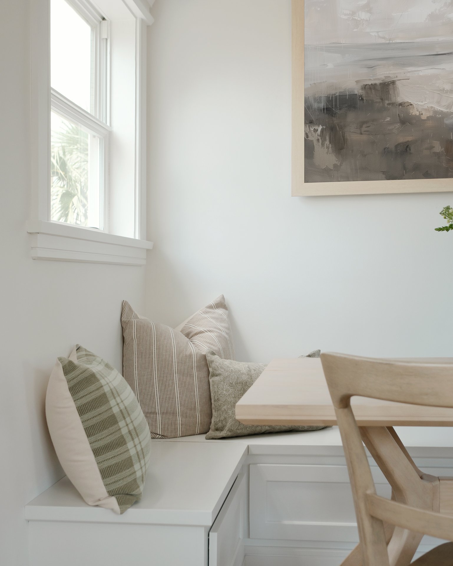 Corner reading nook with white built-in bench, striped and patterned pillows, a window with white trim, and a large framed abstract painting on the wall.