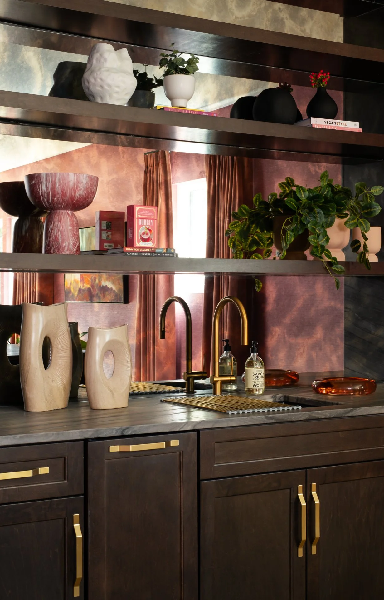 A modern kitchen with dark wood cabinetry, gold hardware, a marble countertop, and gold-tone faucets. The backsplash features a reddish-brown textured wall. Decor includes wooden vases, plants, and shelves with books and vases, with a window and curt