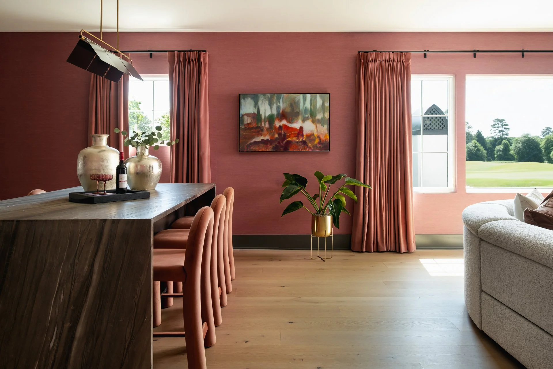 Interior view of a dining area with a wooden table, salmon-colored chairs, pink curtains, a large white sofa, potted plant, wall art, and two windows showing a view of a green landscape.