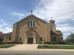 Exterior of a church with a cross on top, beige walls, and a small bell tower, under a cloudy sky.