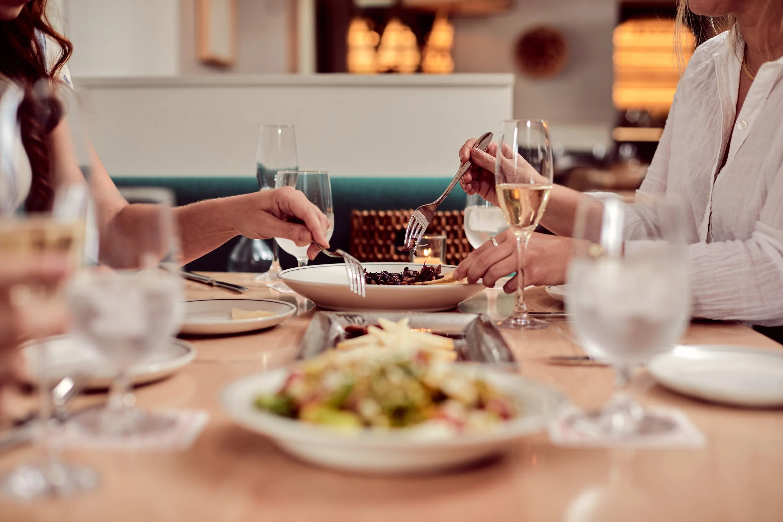 People dining at a restaurant table with wine glasses and plates of food, including salad and a pasta dish. Hands are holding forks.