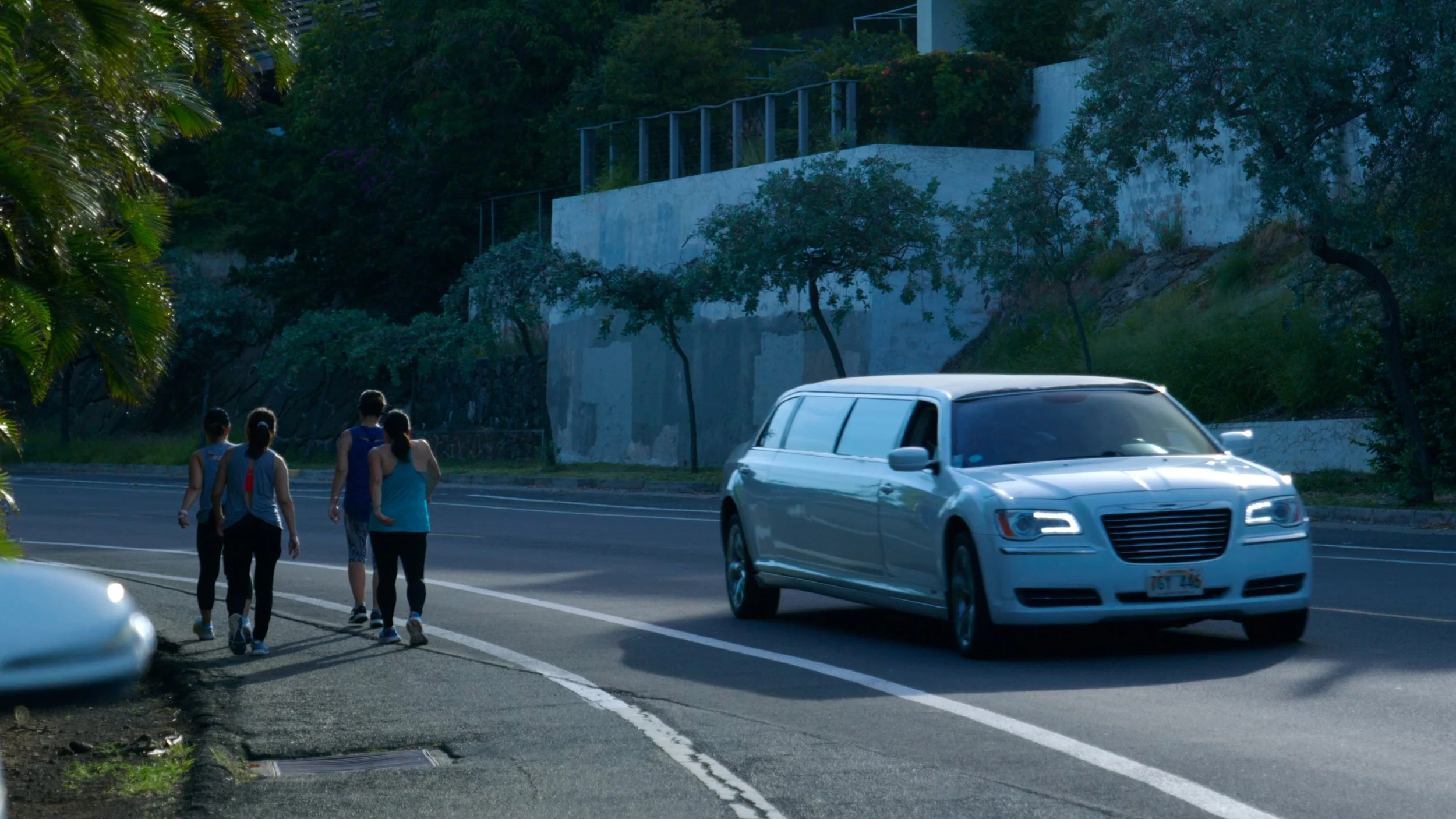 Limousine driving past pedestrians on a tree-lined street. Still from Priced Out of Paradise, a Hawaii housing documentary directed by Annie and Sam Menendez
