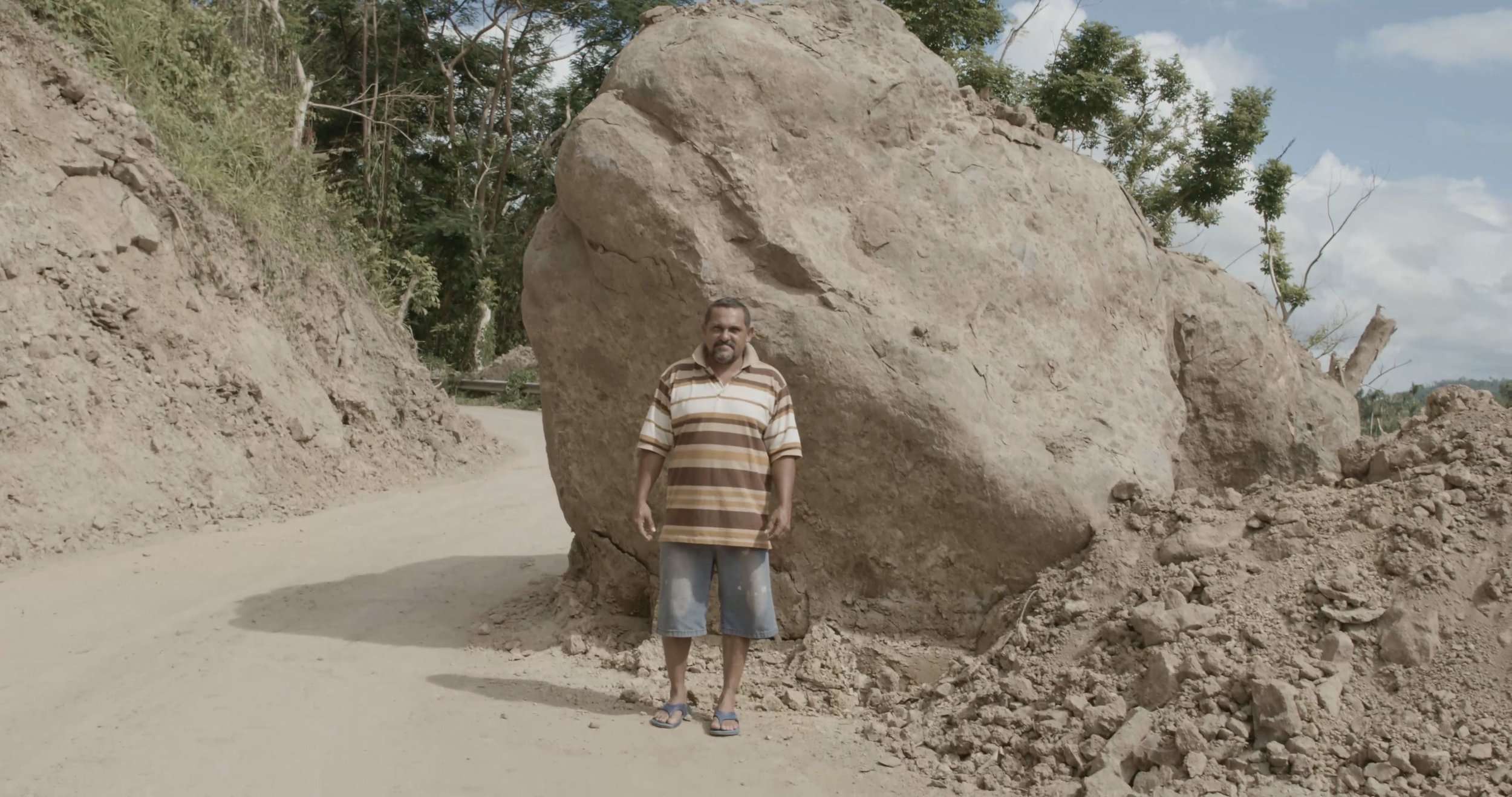 Enrique Lorenzo beside a giant boulder blocking a road in Utuado, Puerto Rico, after Hurricane Maria. Still from Hurricane Maria: Stories from the Storm, directed by Sam Menendez and produced by Annie Menendez