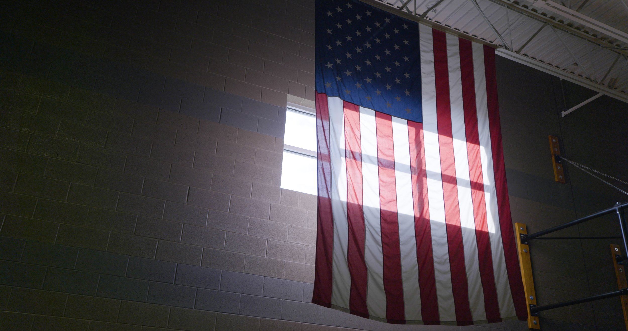 American flag lit by window light in a dark room. Still from The Real Cost, a student debt documentary directed by Sam Menendez and produced by Annie Menendez