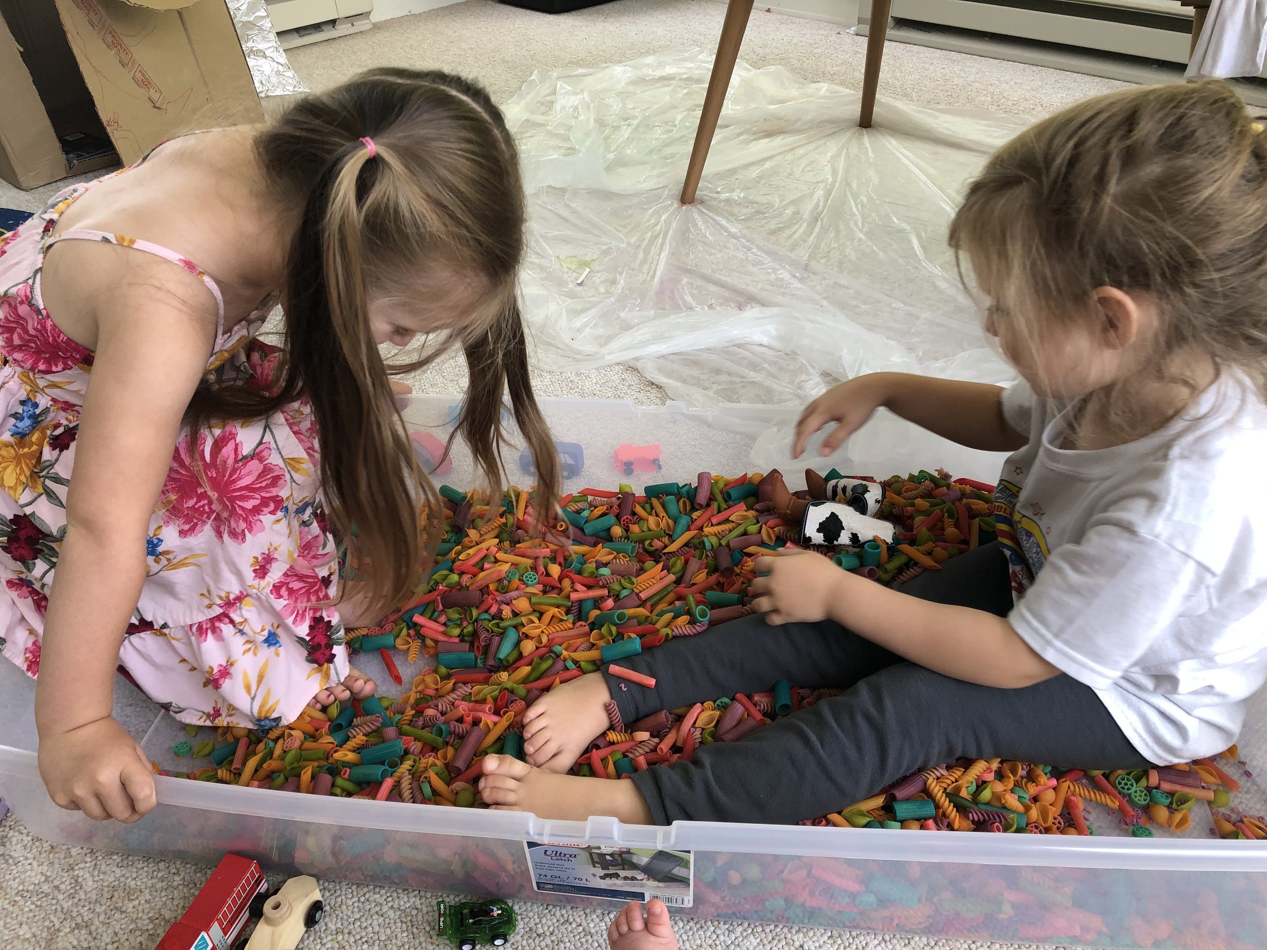 Two children sitting in a sensory bin full of rainbow pasta