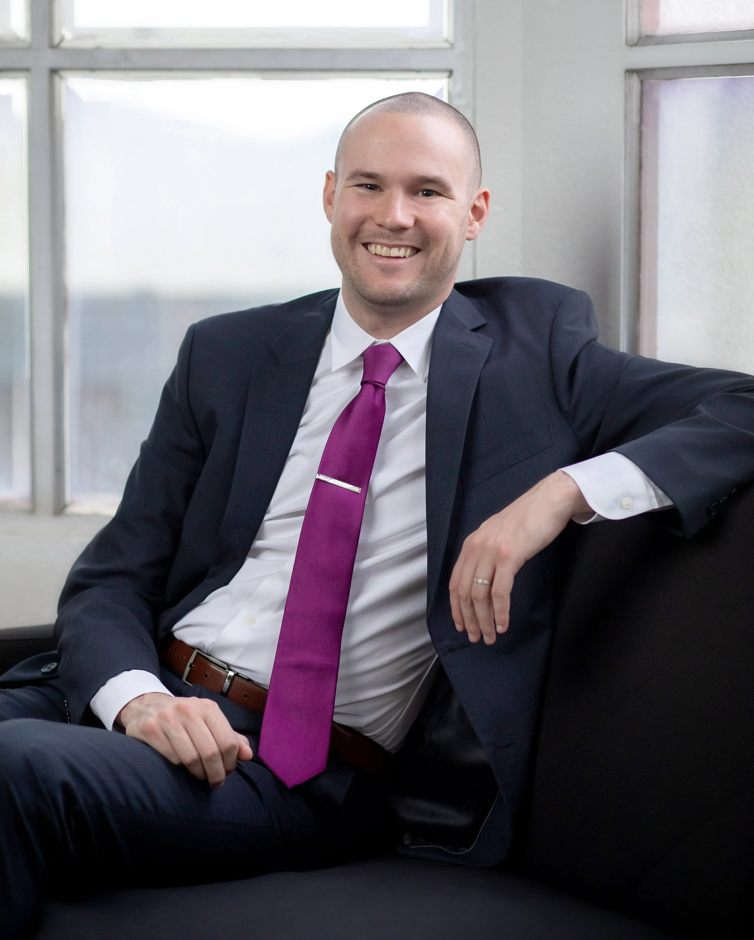 Chris wearing a black suit, white shirt, and magenta tie, sitting on a sofa.
