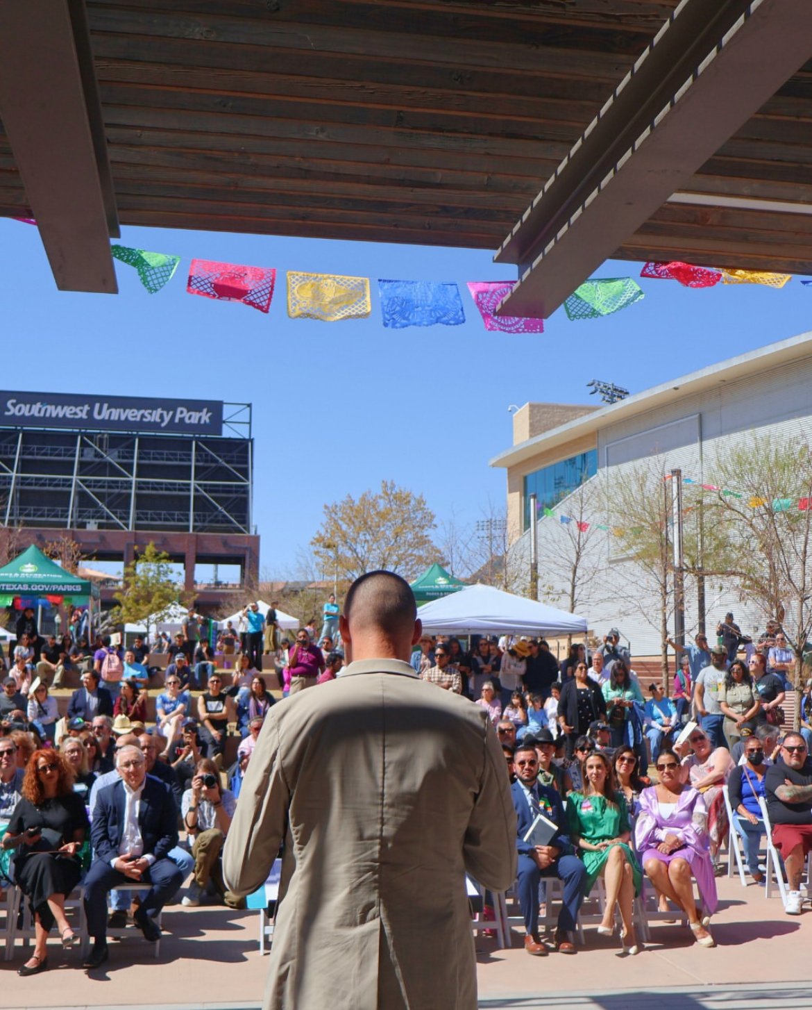 Chris addressing a large crowd seated in Cleveland Square Park at the grand opening of the Mexican American Cultural Center in Downtown El Paso. The crowd is seated in tiered seating with tents and colorful decorations.
