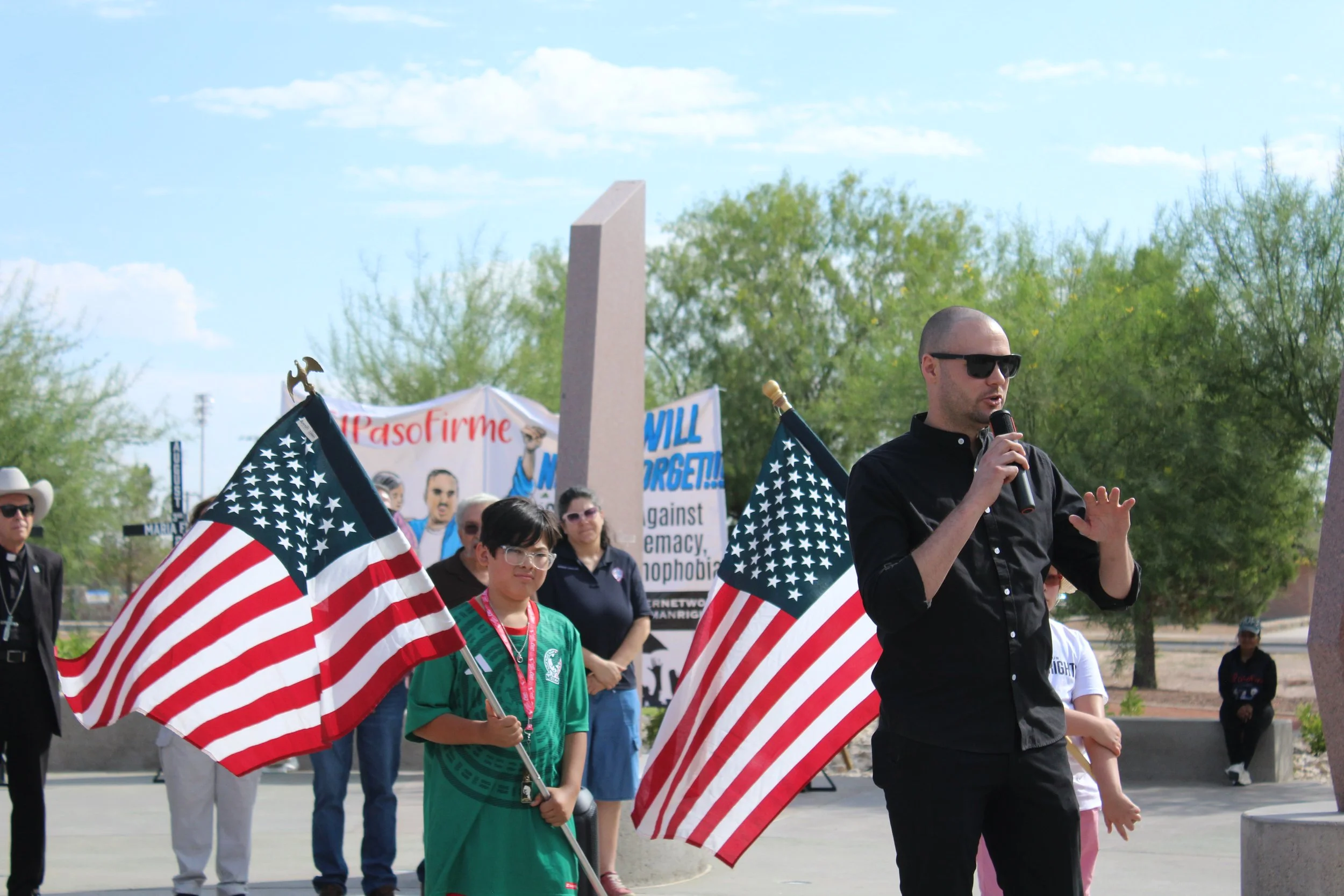 Chris wearing sunglasses and a black shirt speaking into a microphone at a Border Network for Human Rights event, surrounded by people holding American flags and signs, with trees and a blue sky in the background.