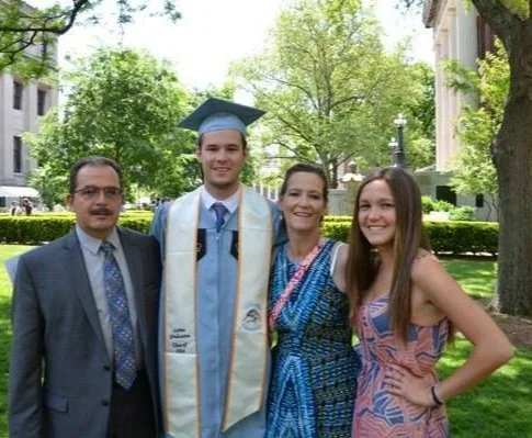Chris, at his graduation from Columbia University, in graduation cap and gown with his mother, father, and sister.