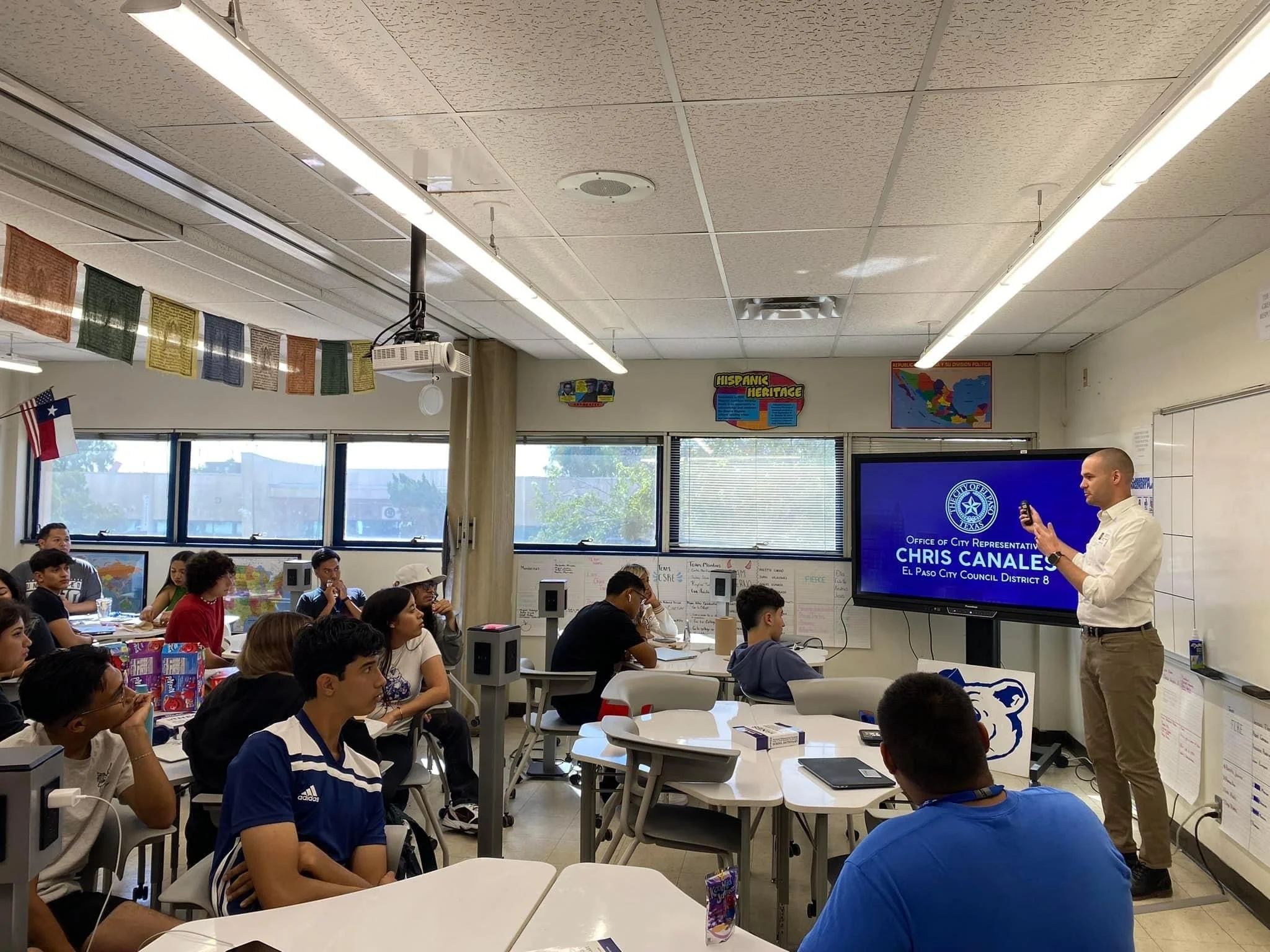A government class at Bowie High School filled with students seated at desks, attentively listening to a Chris, who is standing in front of a large screen and delivering a presentation about local civics.