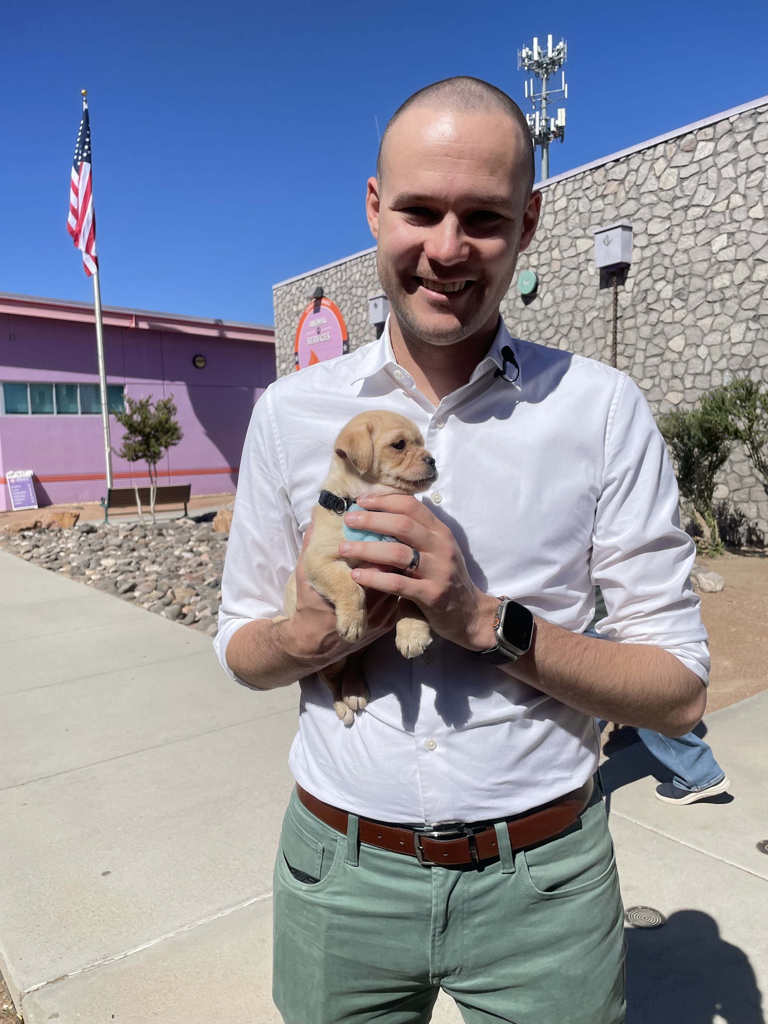 Chris wearing a white shirt and green trousers holding a tiny puppy, standing outdoors at the El Paso Animal Services shelter.