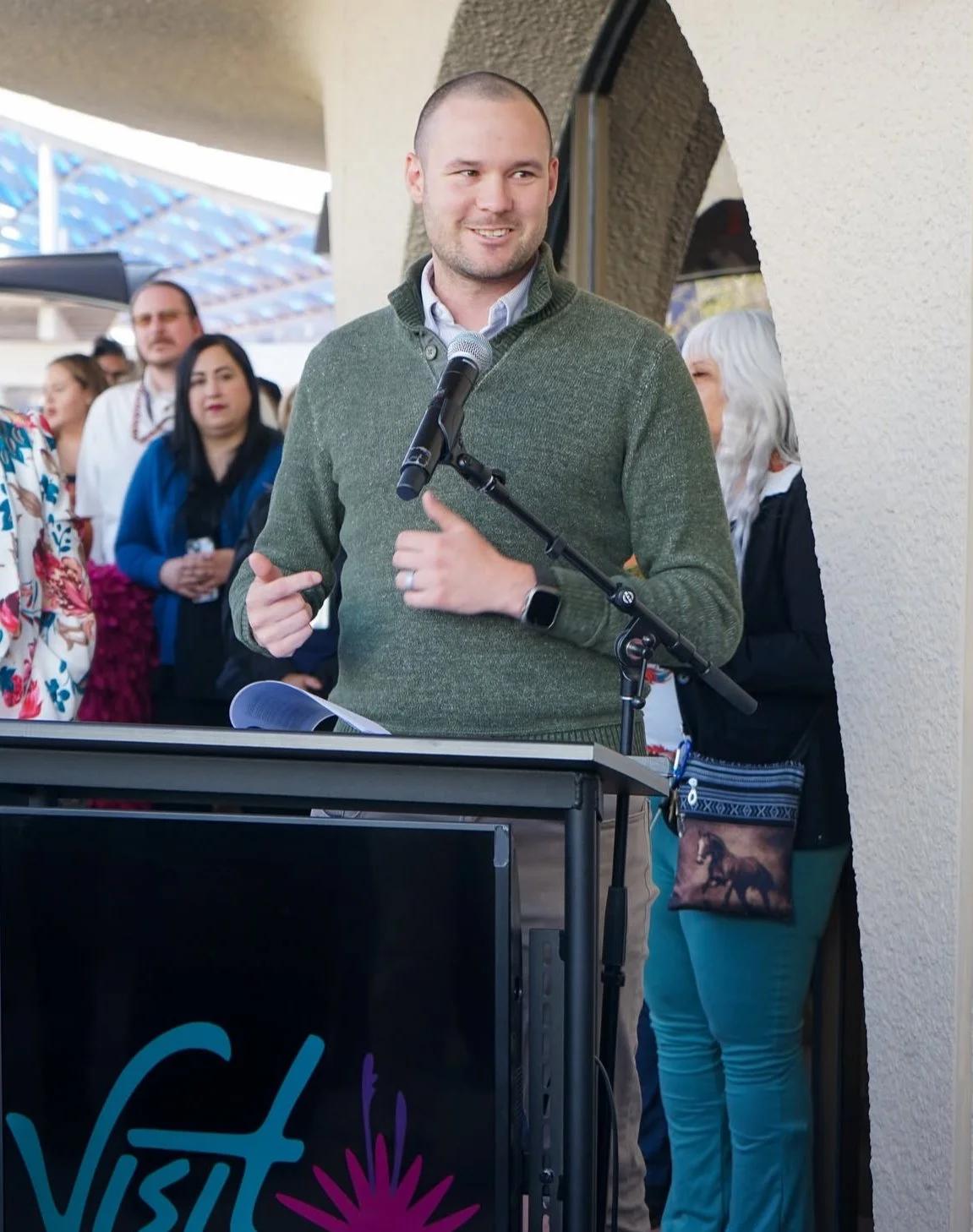 Chris speaking at a podium with a microphone at an outdoor event at the Downtown Visitor's Center, surrounded by a group of people.