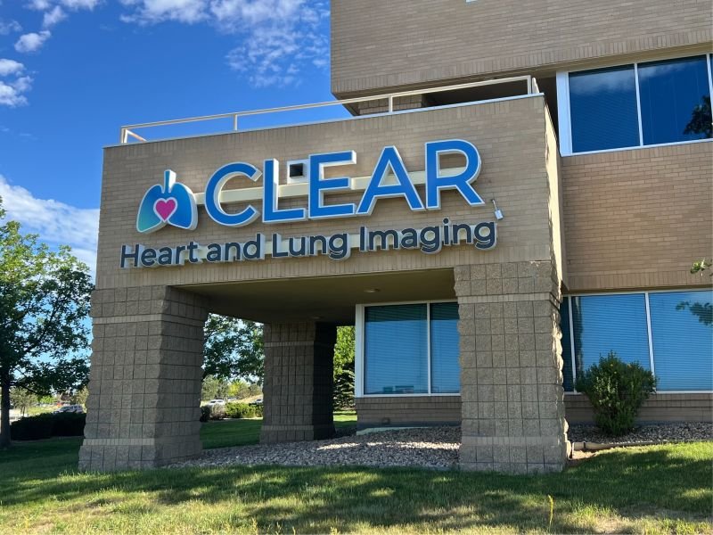 Exterior of a medical facility with a sign that reads 'CLEAR Heart and Lung Imaging,' featuring a logo of a heart inside a stylized image of lungs, on a sunny day with a clear blue sky.