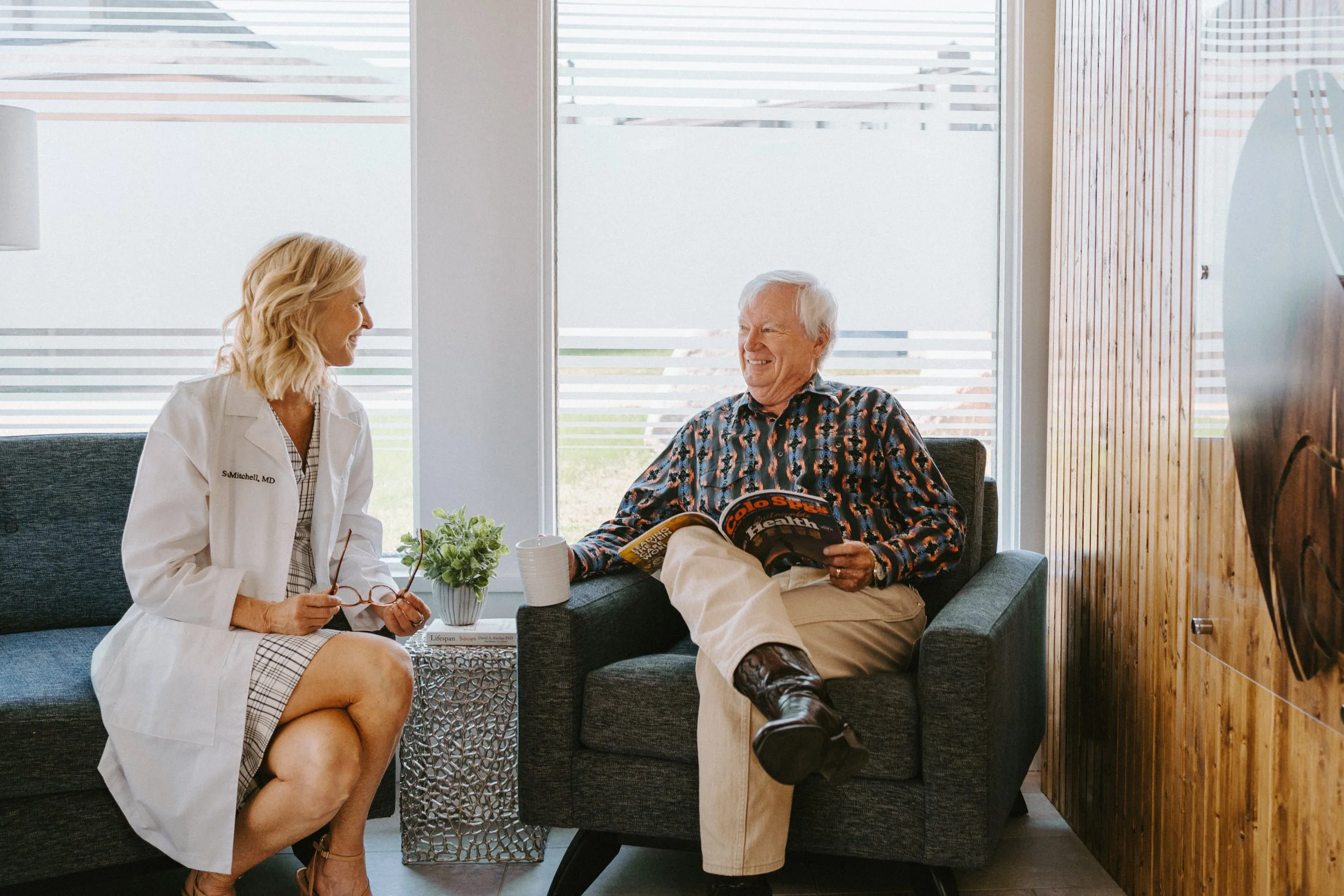 A female doctor and an elderly man are sitting in a waiting room, engaged in conversation and smiling. The woman is holding glasses, and the man is reading a magazine. There is a plant on a small table between them and a large window in the background.