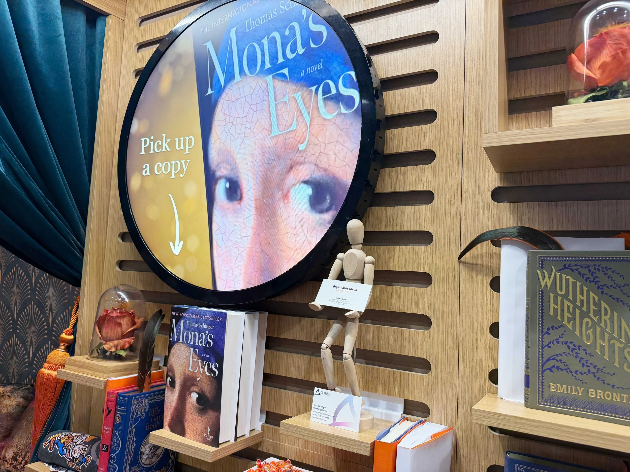 Round circular digital signage display showcasing "Mona's Eyes" graphics next to books on a shelf, highlighting the multi-vendor compatibility challenge.