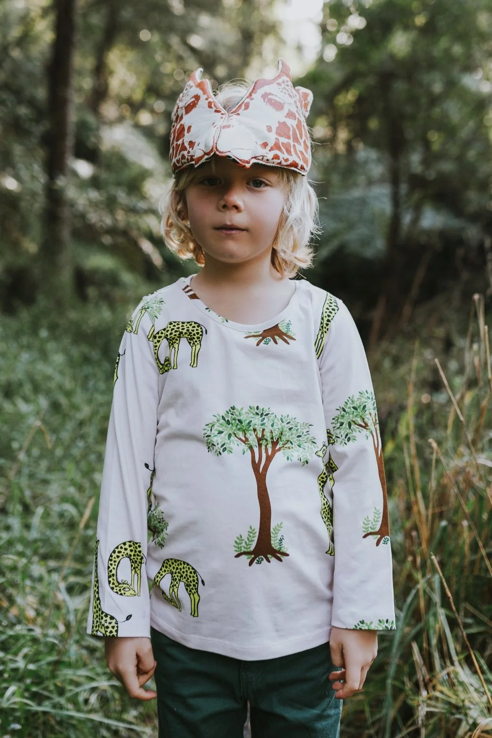 Boy stands wearing giraffe mask in a field for kids clothing photo shoot.