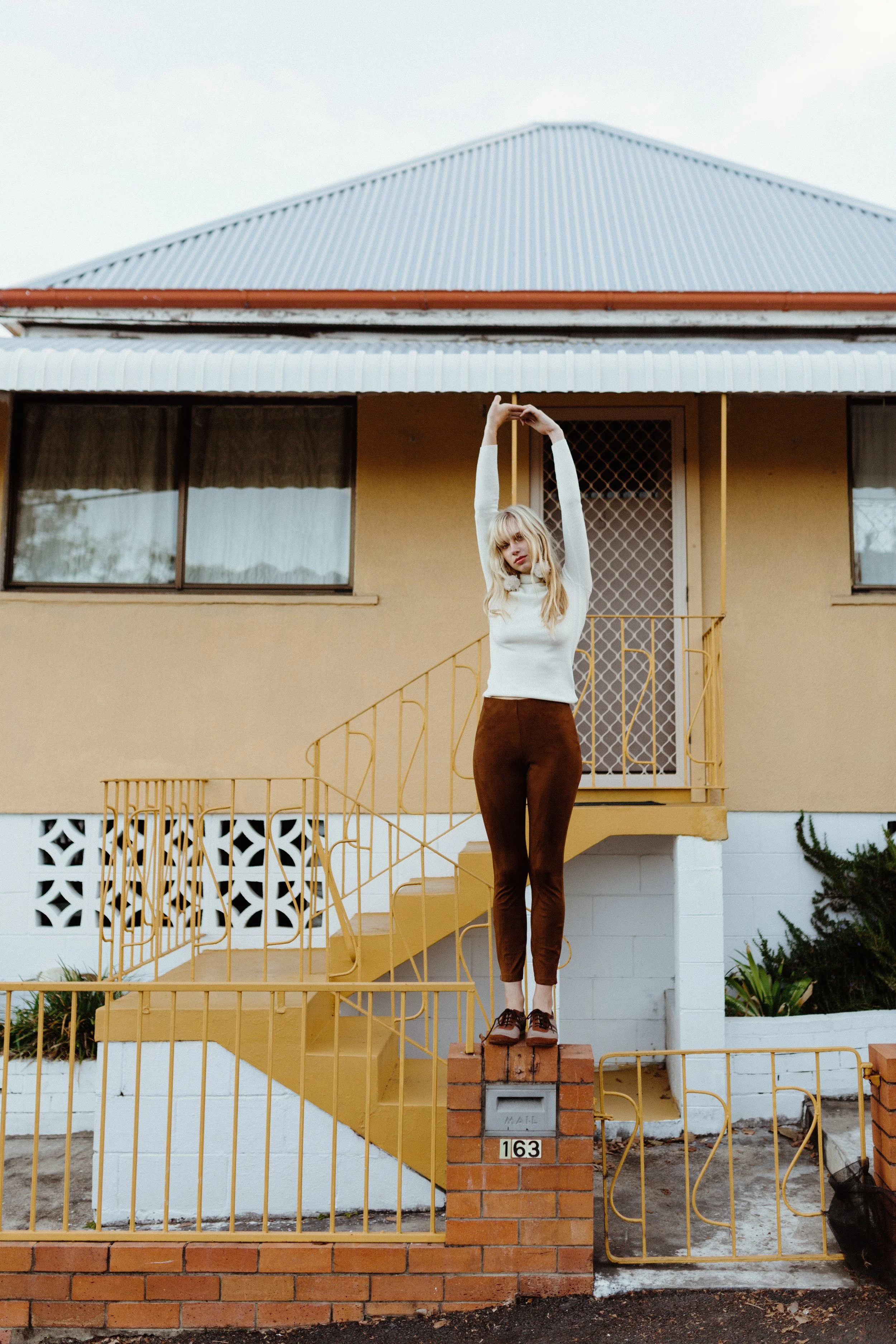 Model stands on fence in front of retro house yellow house in Brisbane, Qld.