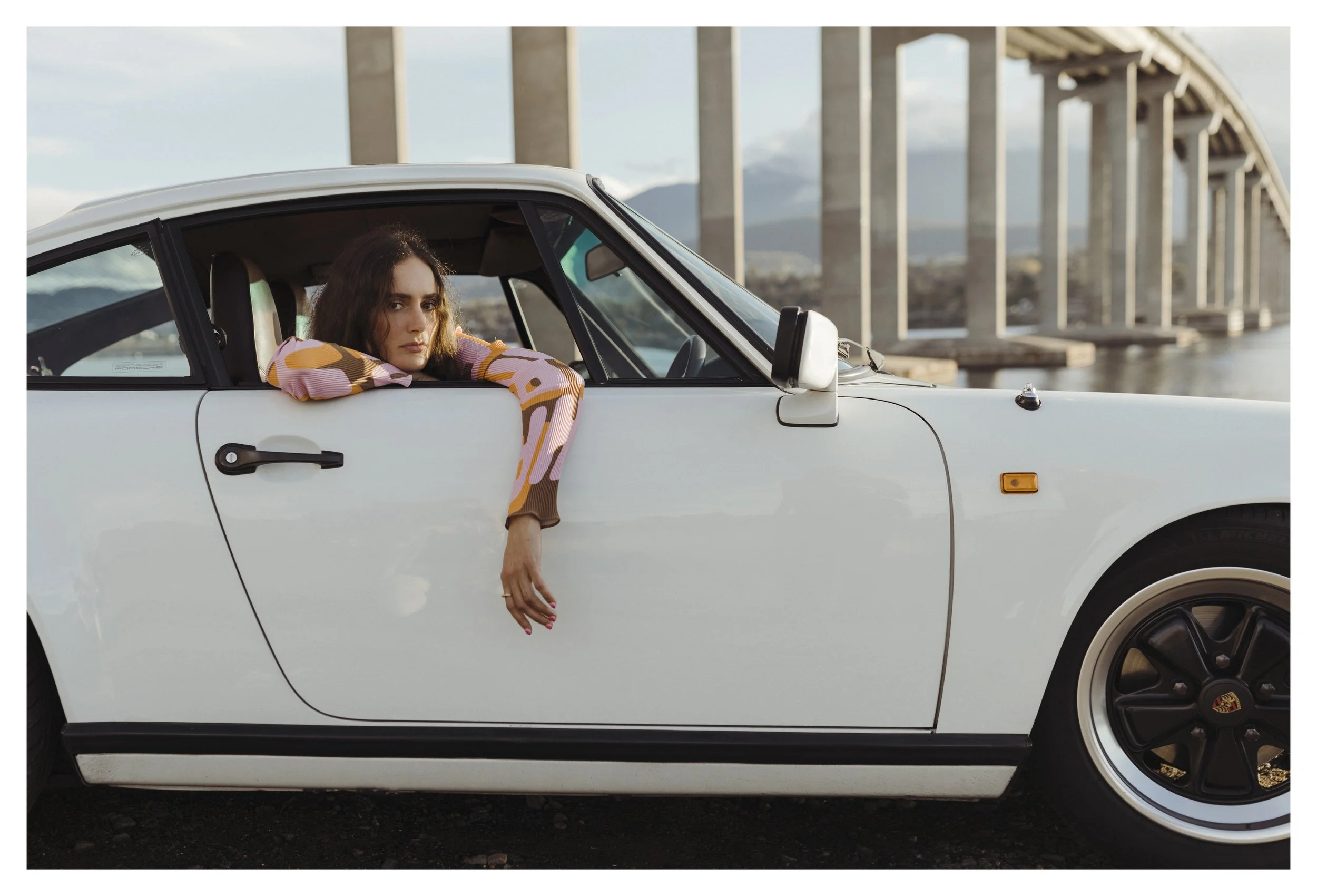 Model poses in vintage car next to a bridge.