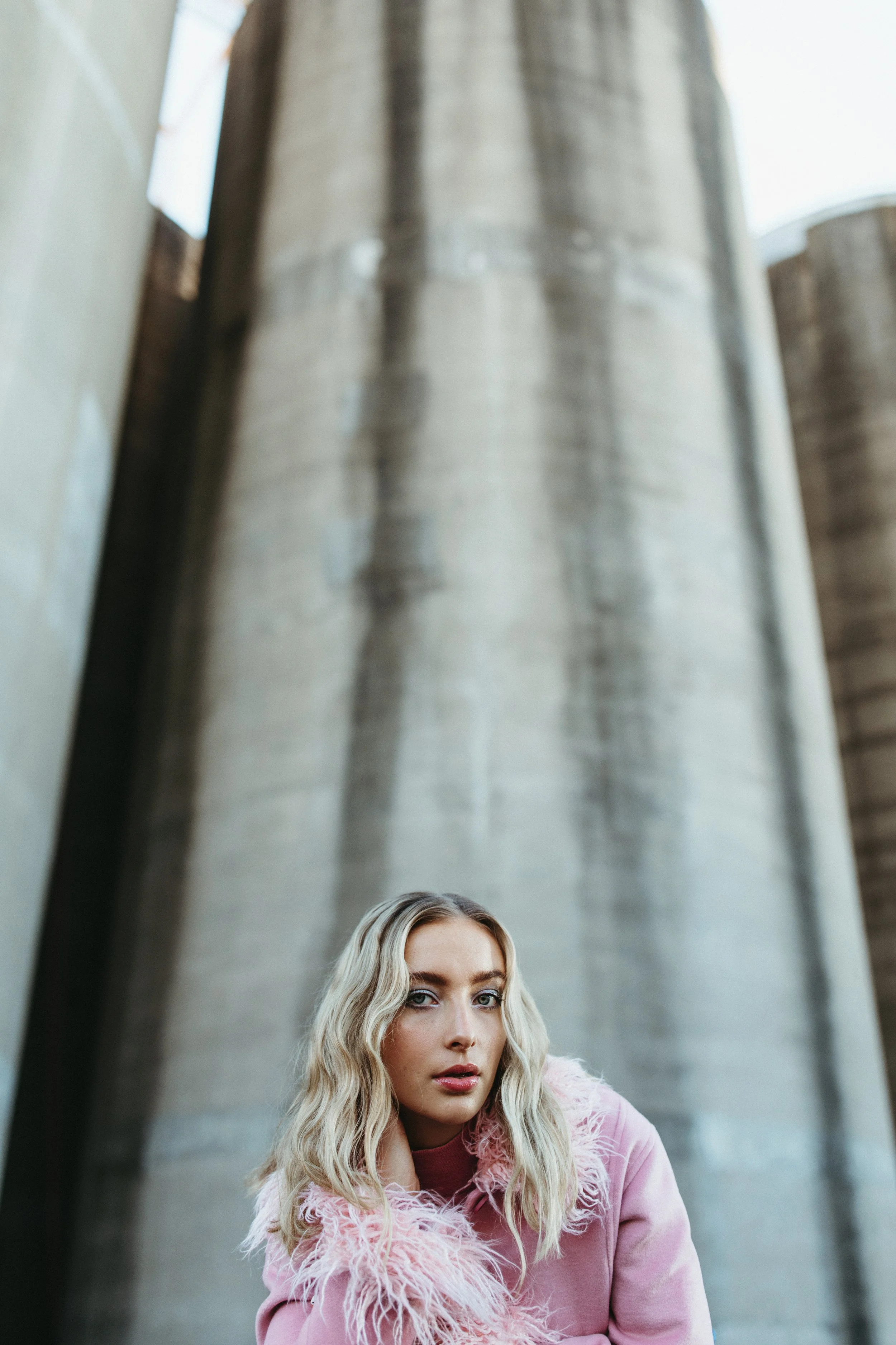 Model in pink jacket poses in front of silos for fashion shoot.