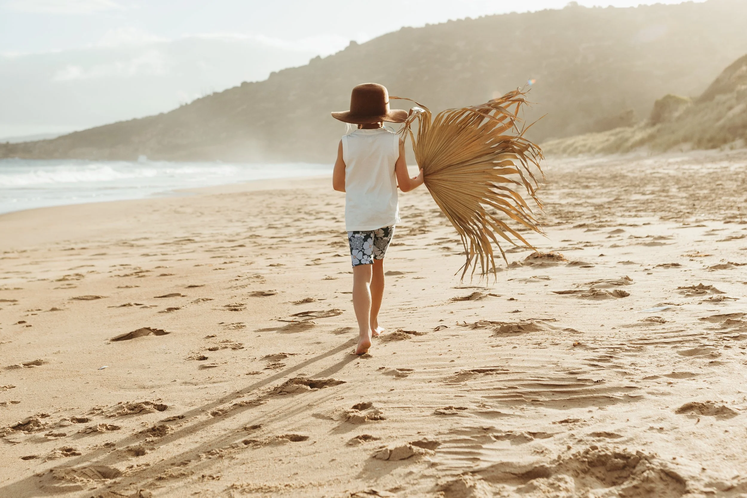 Girl walks along beach with palm frond during kids fashion photo shoot.