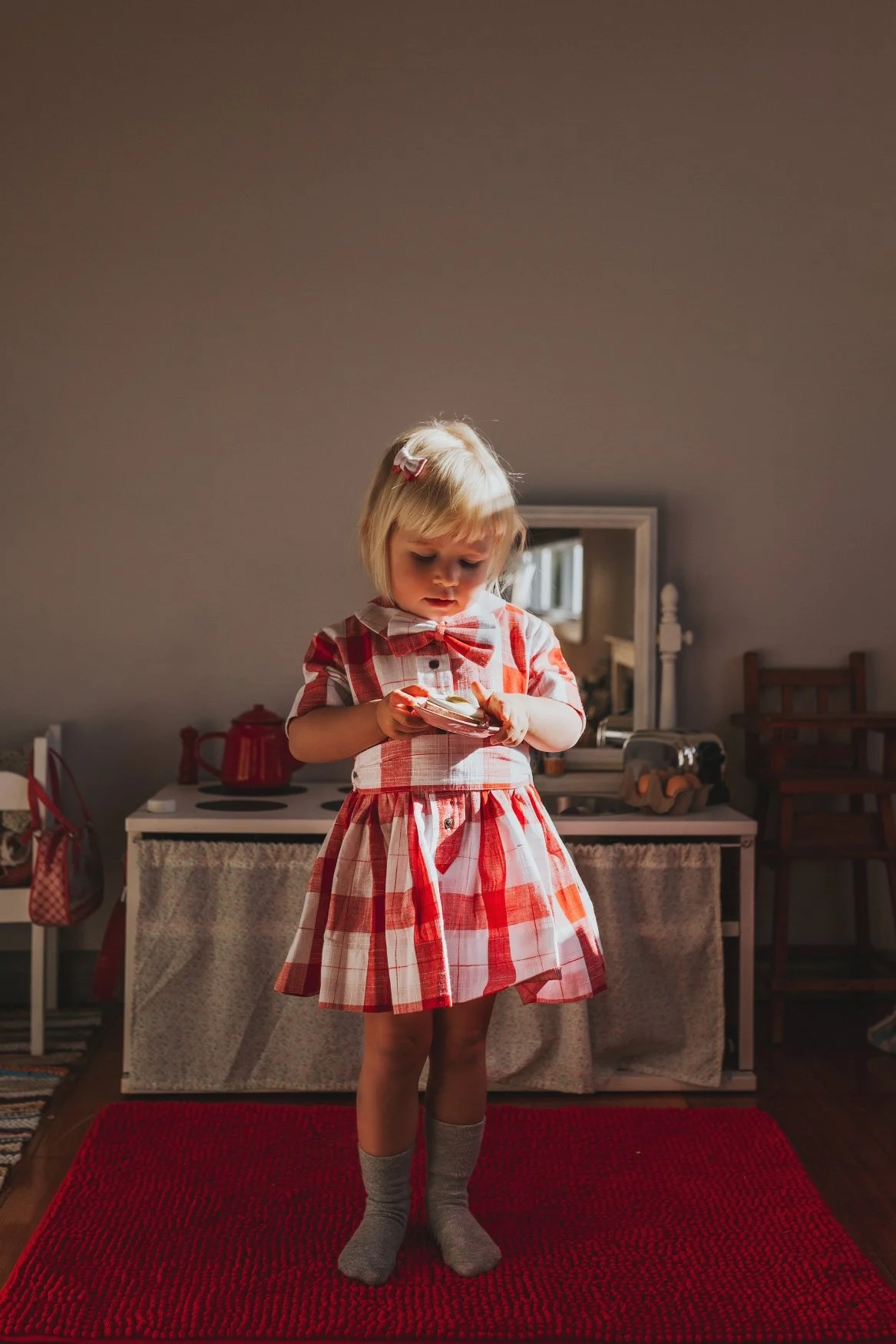 Young girl stands in red check dress in sunny spot of her house.