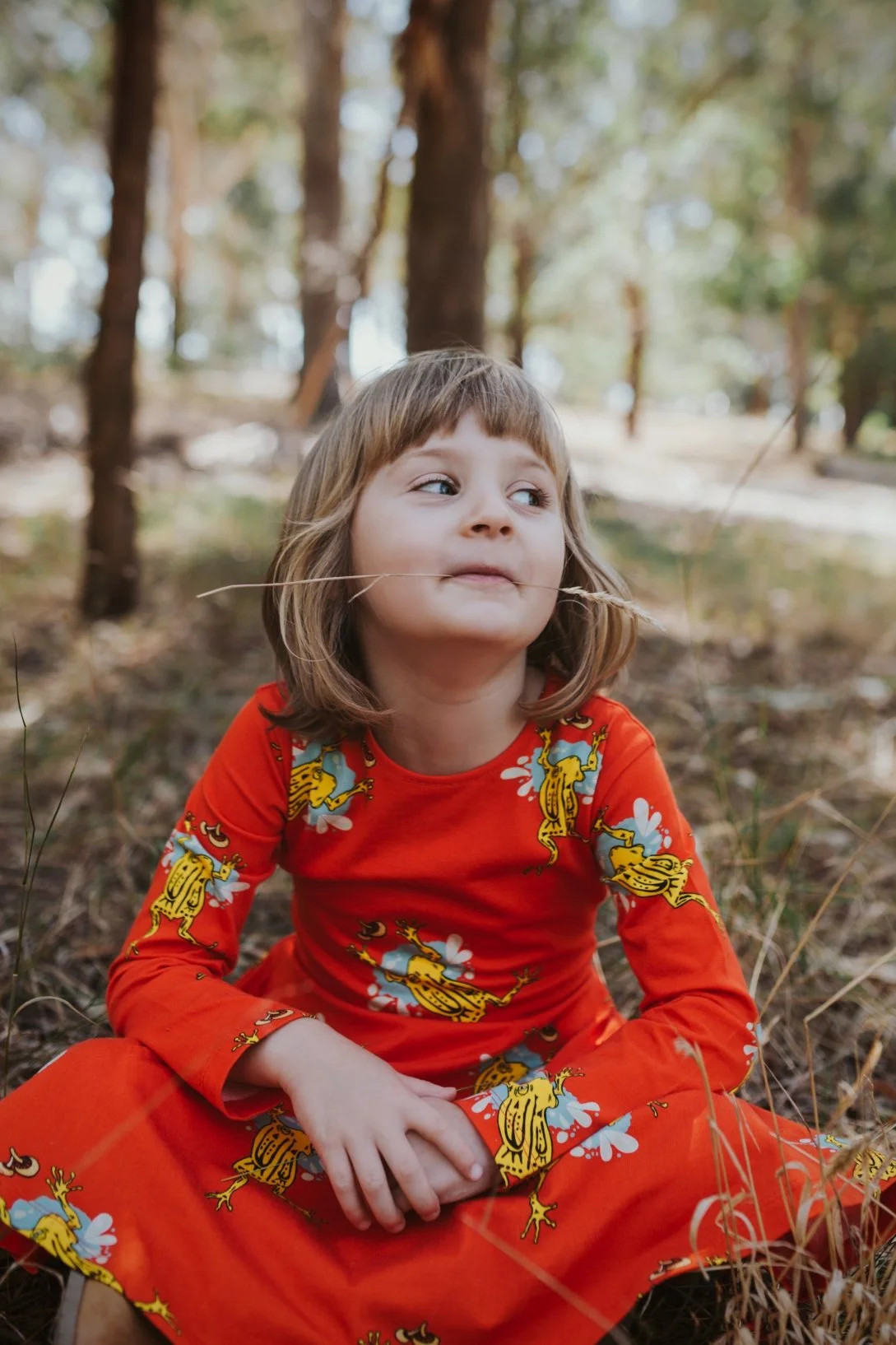 Young girl sits on grass with dry blade of grass in her mouth.