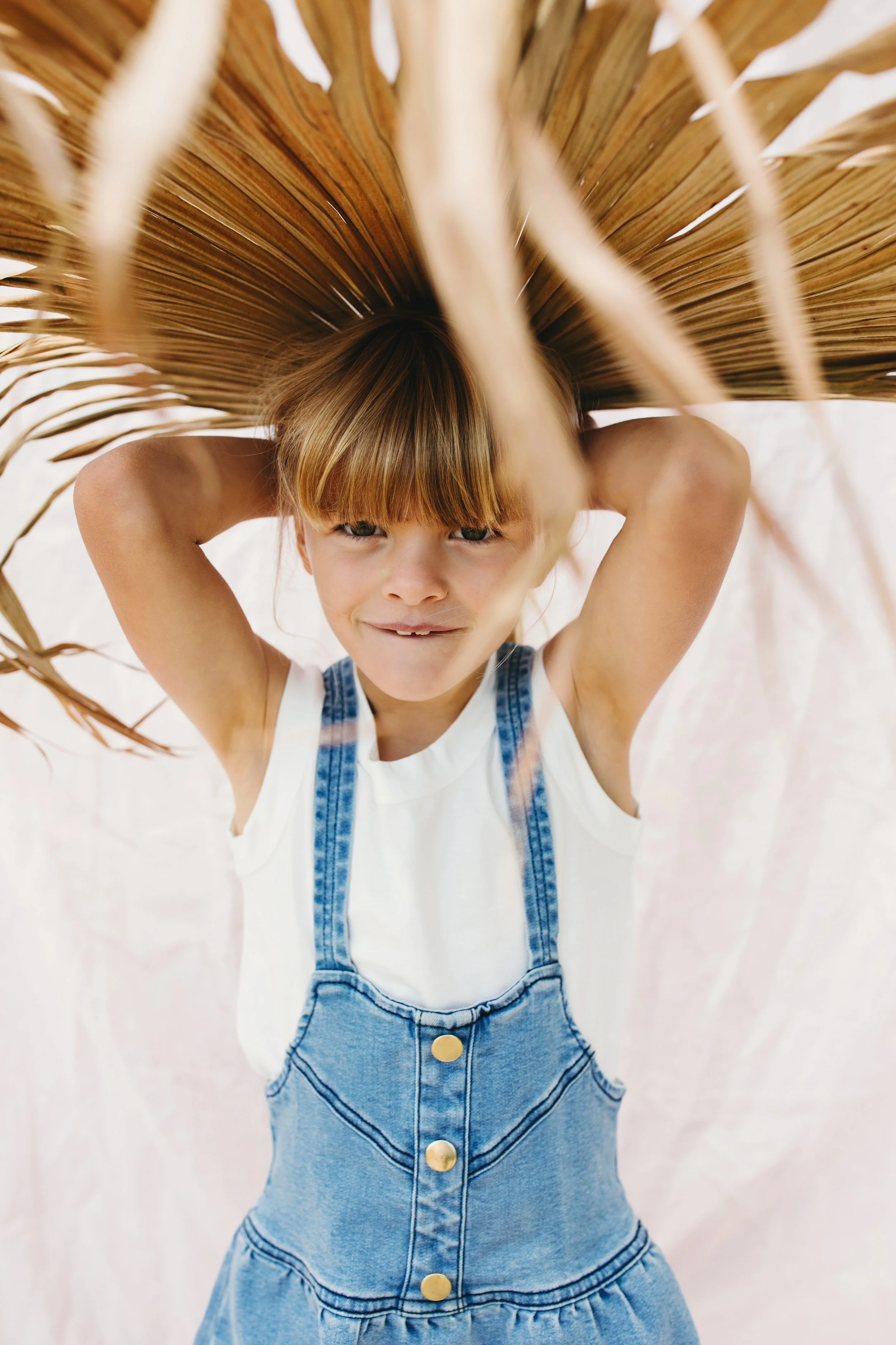 Young girl wears denim dress holding palm frond in studio photo shoot.