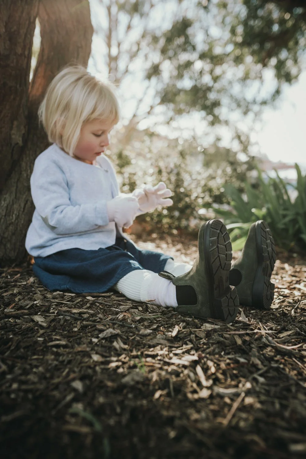 Child sits under tree wearing boots for commercial fashion shoot.