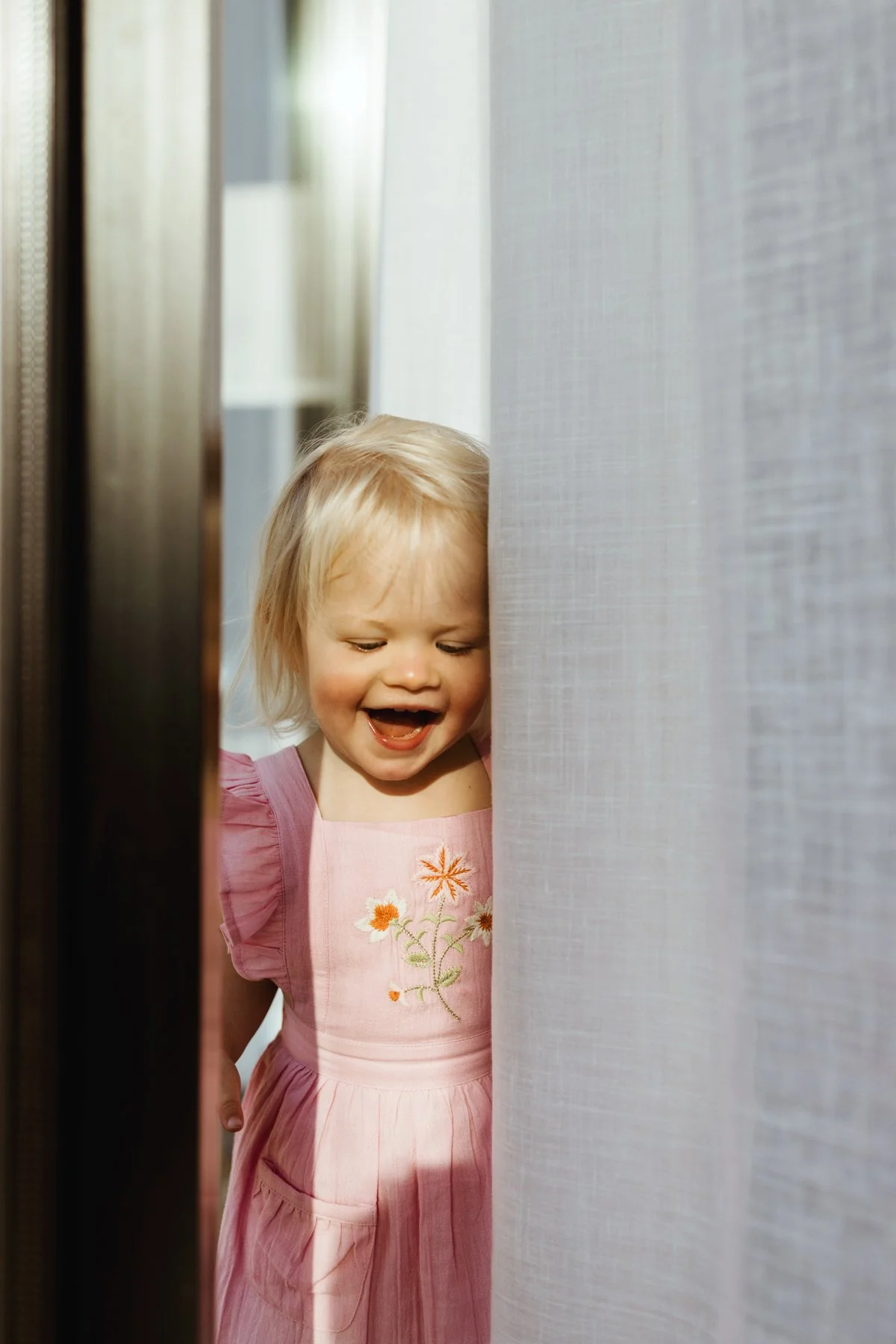 Girl smiles from behind curtains wearing pink dress.