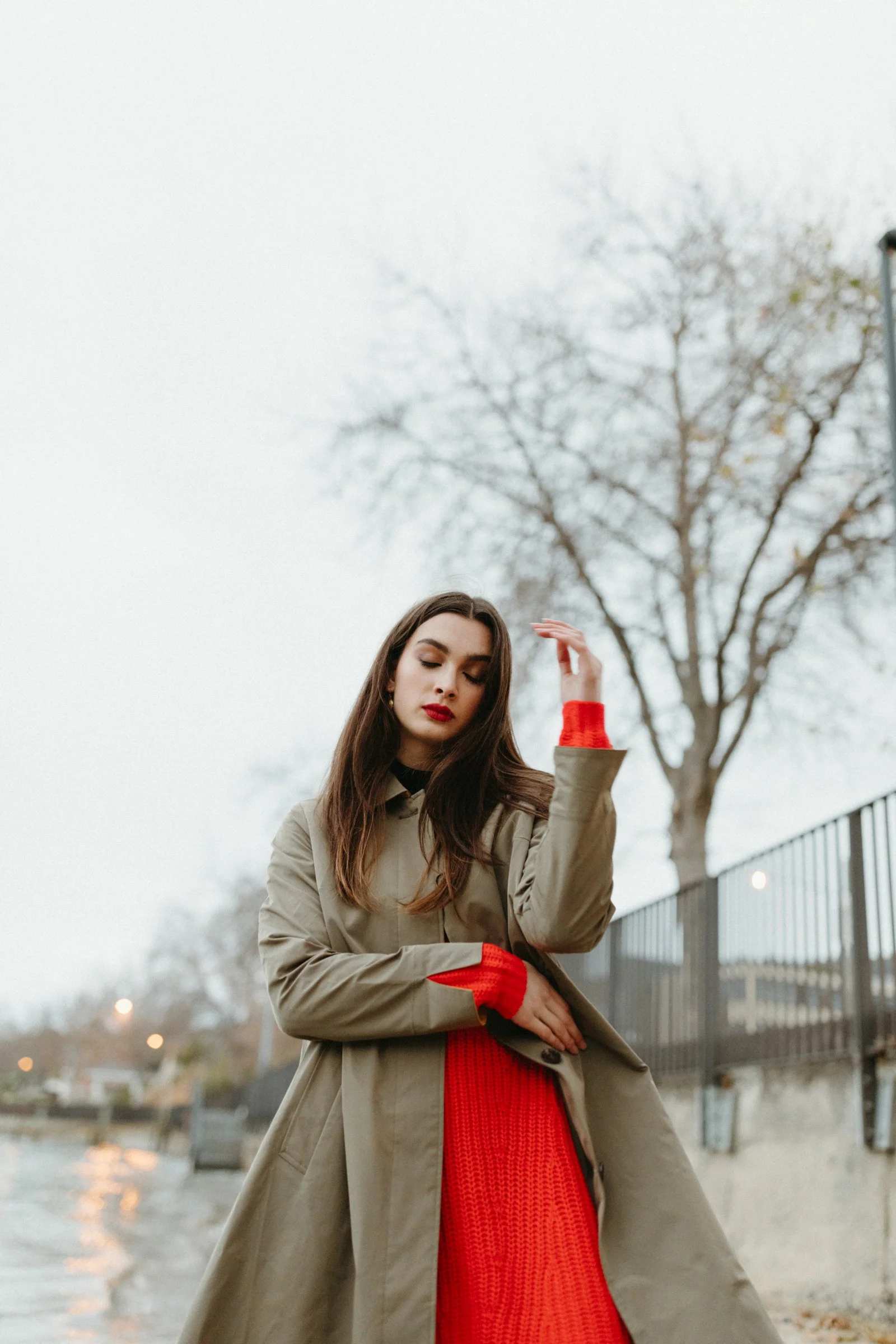 Woman stands with eyes closed in red sweater and trench coat.