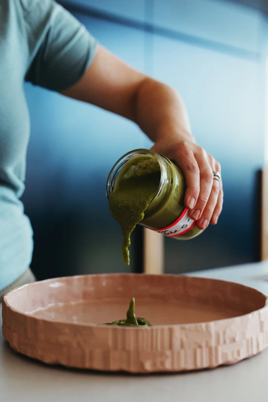 Food writer pours condiment into pink serving dish during recipe book shoot.