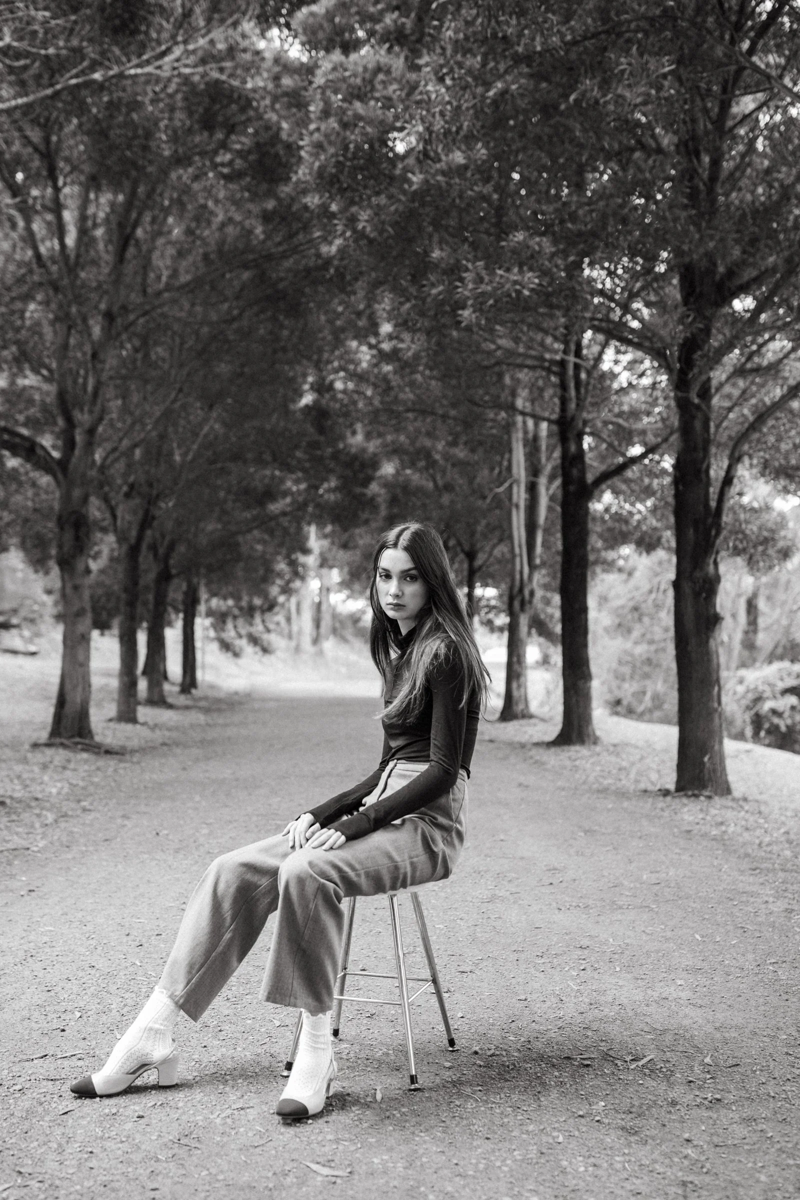 Woman with dark hair sits on stool on pathway through park.