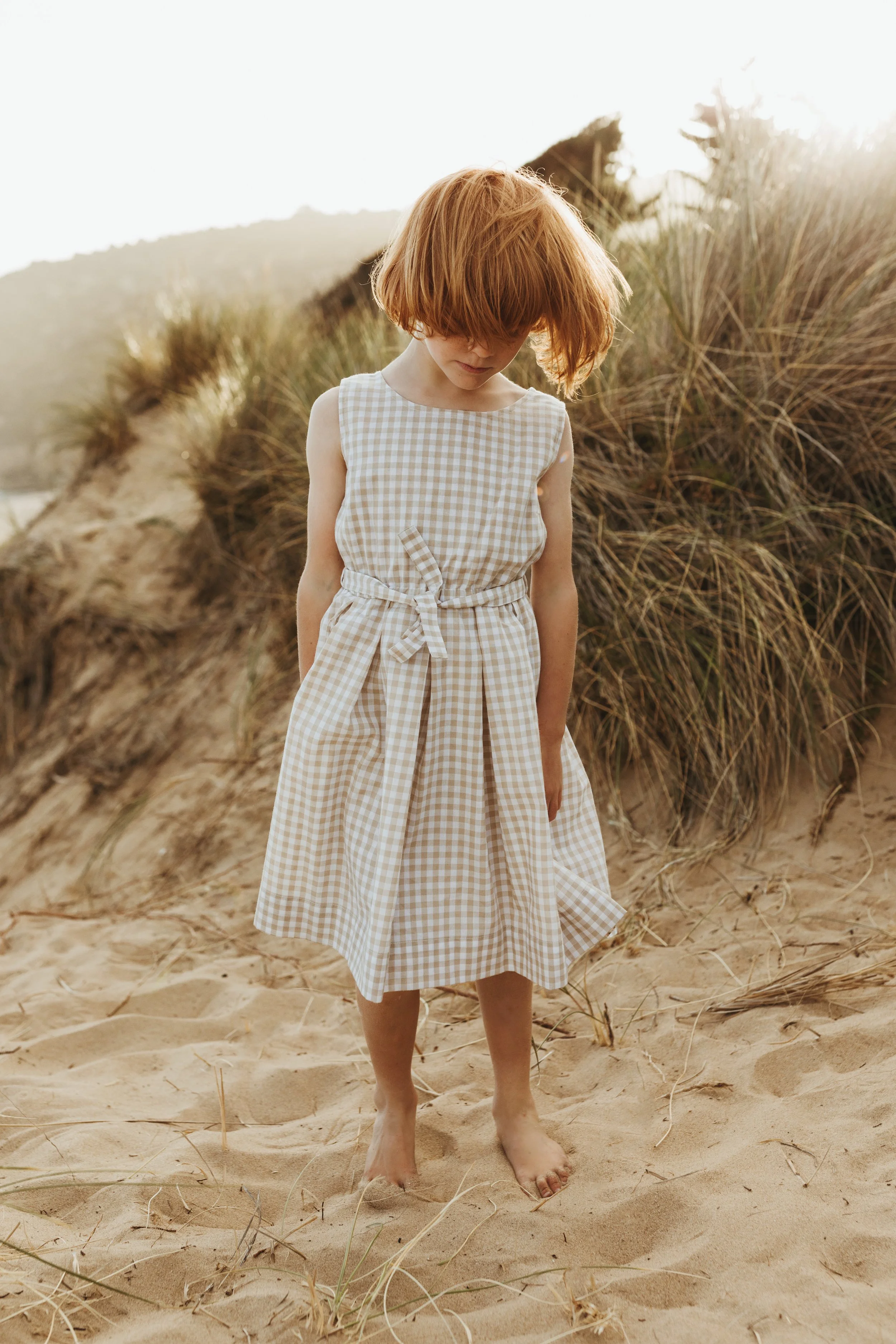 Girl stands on beach in sweet gingham dress for kids label shoot.