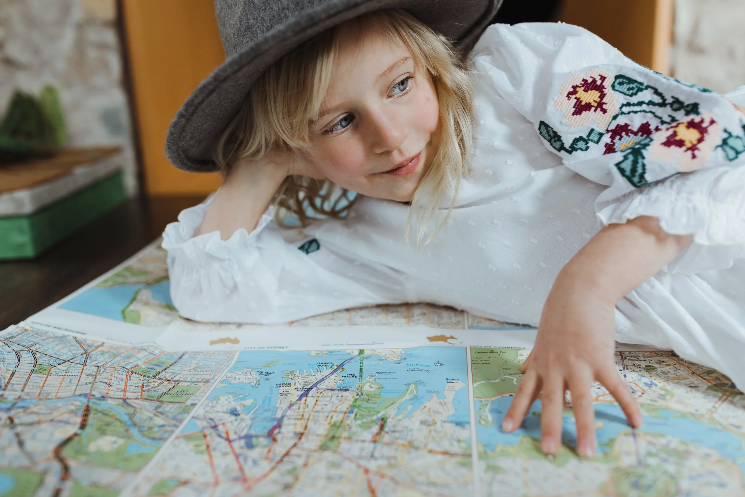 Blonde girl lays on map wearing felt hat and white dress.