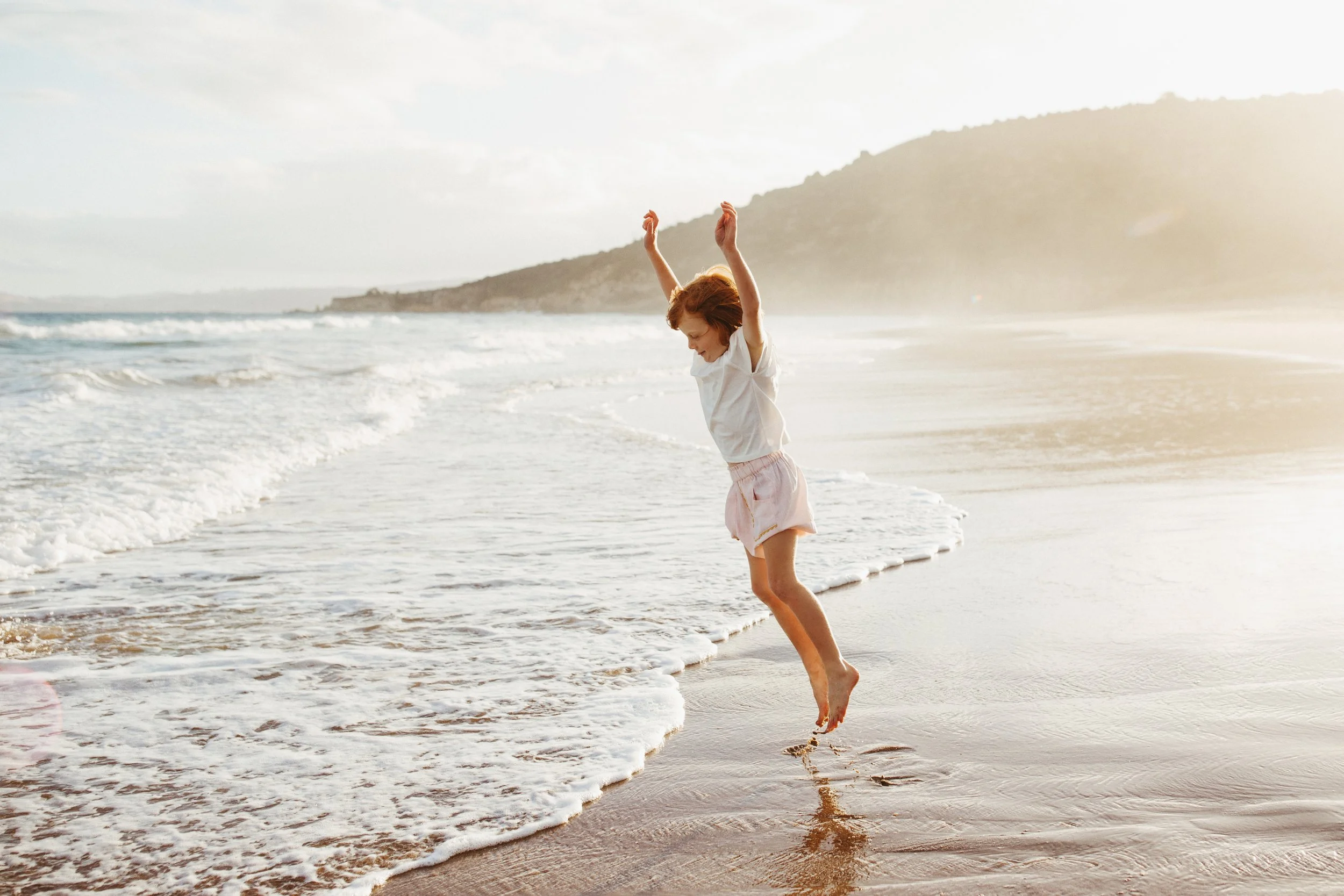 Young girl jumps at water's edge during kids' fashion shoot.